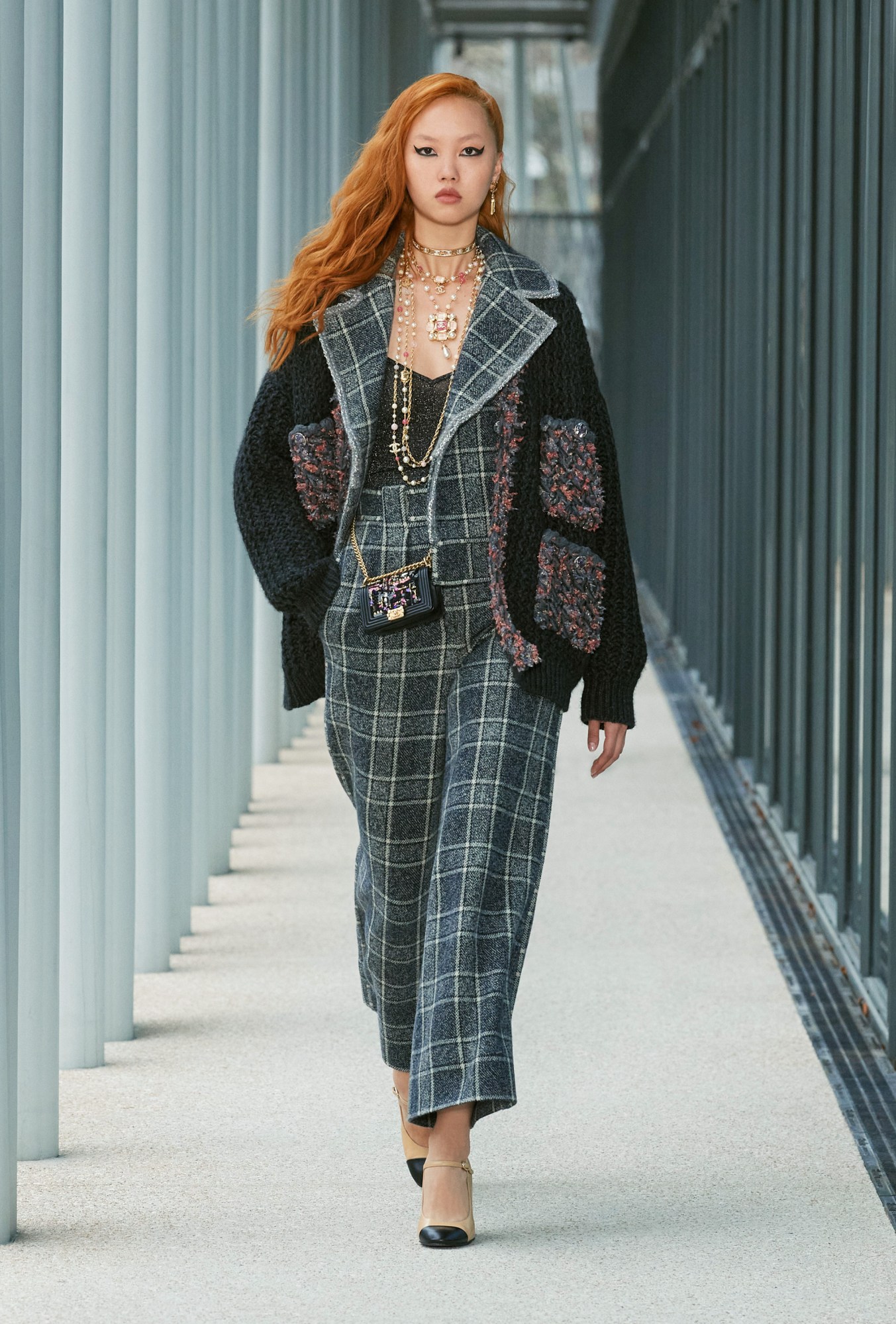 Model wears a two piece check suit with a knitted oversized cardigan and a black handbag and lots of necklaces.