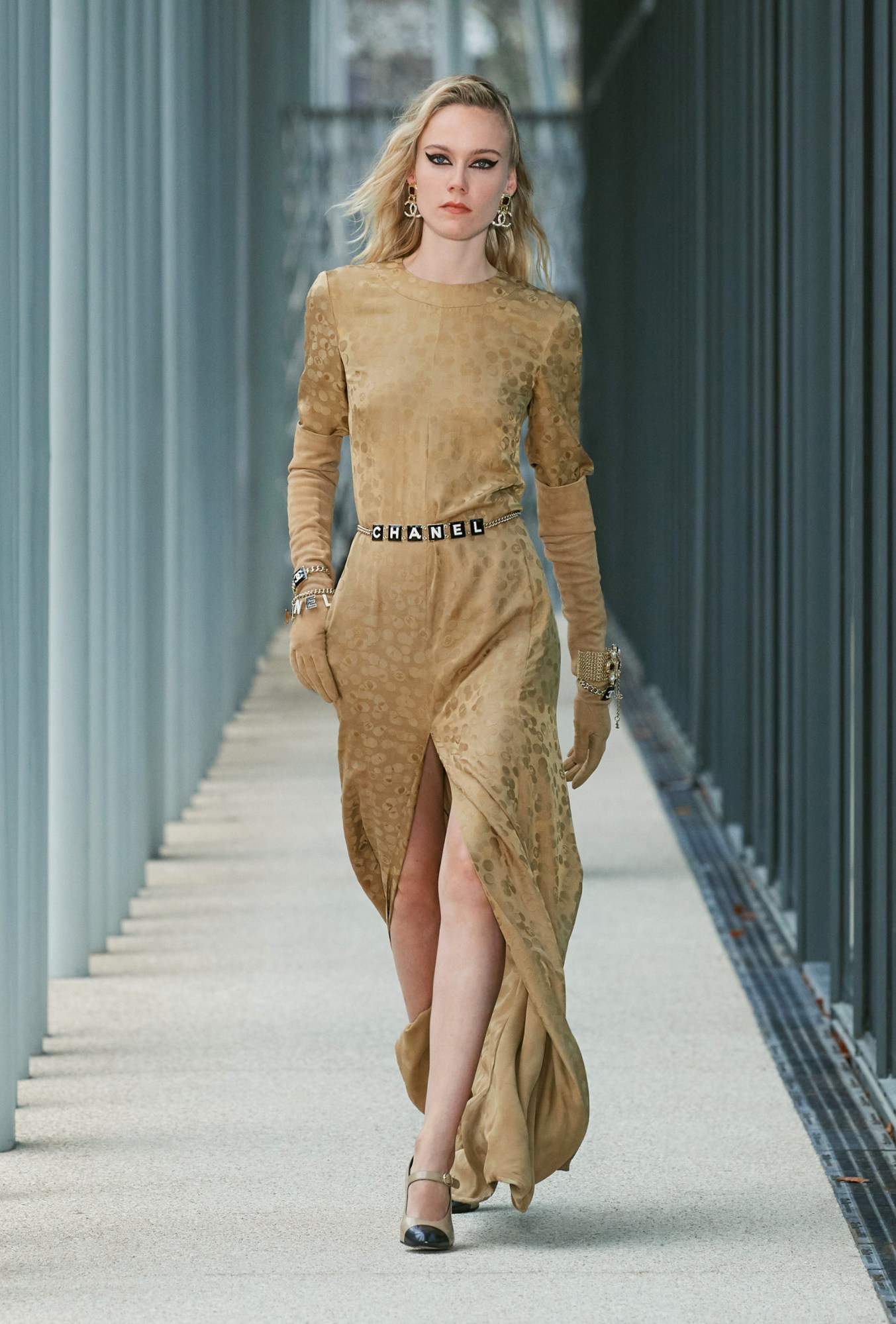 Model wears a gold printed high neck dress with long sleeves, tan gloves and a Chanel lettering belt.