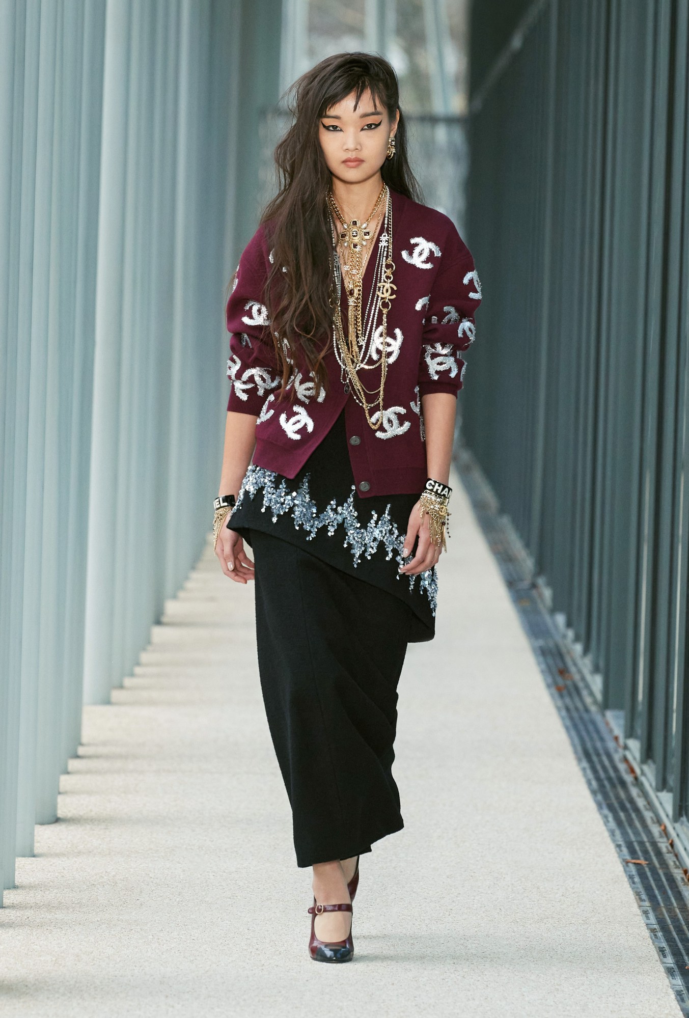 Model wears a black maxi skirt with an embroidered peplum layer and a burgundy cardigan with the Chanel logo printed on it.