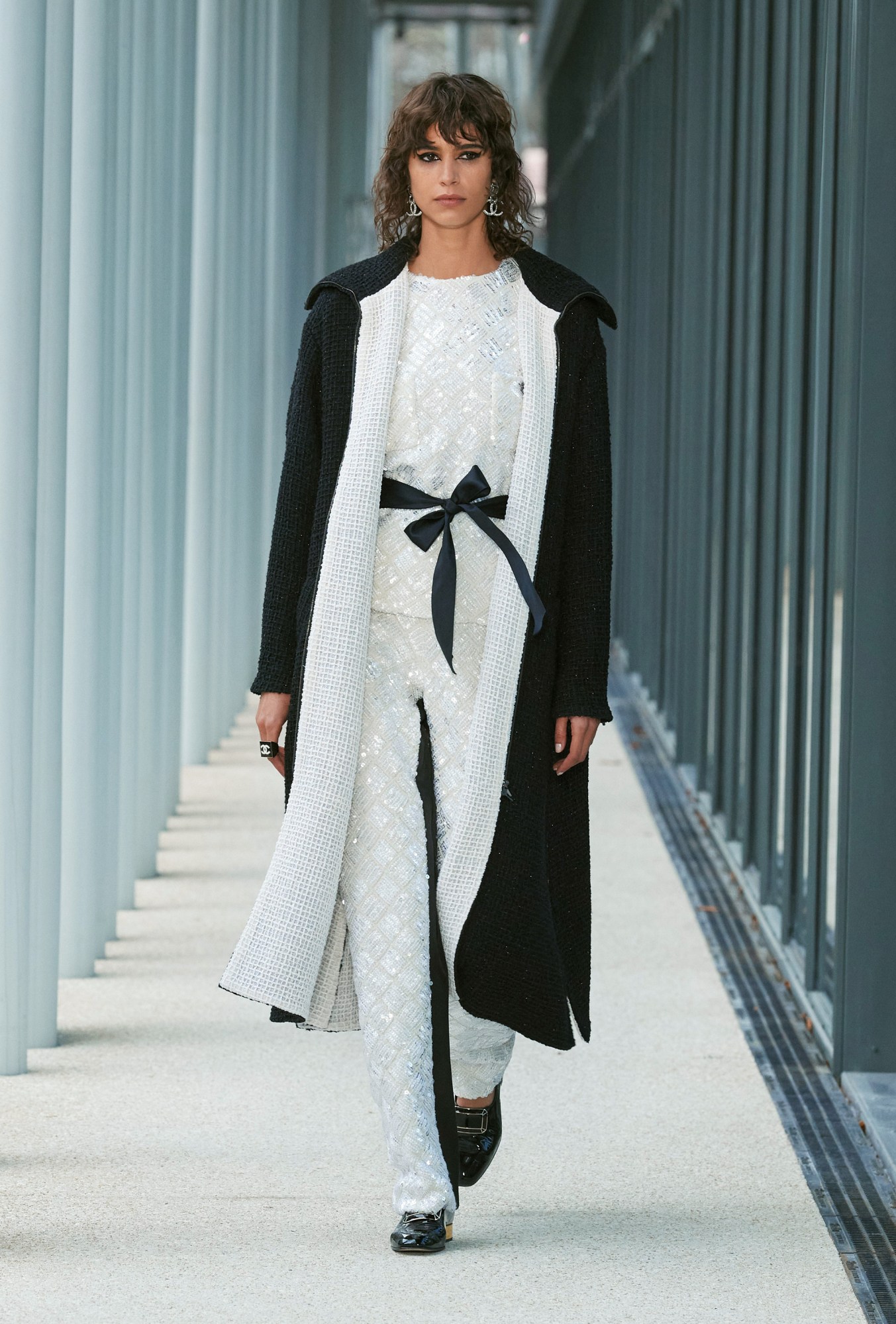 Model wears a white crystal embellished pant suit with a black bow and a woven black overcoat with white lapels and lining.