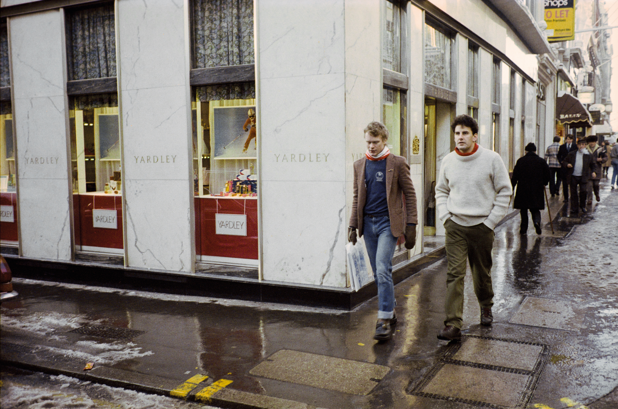 taken by sunil gupta in 80s london, two white men in casual dress walk down a wet pavement