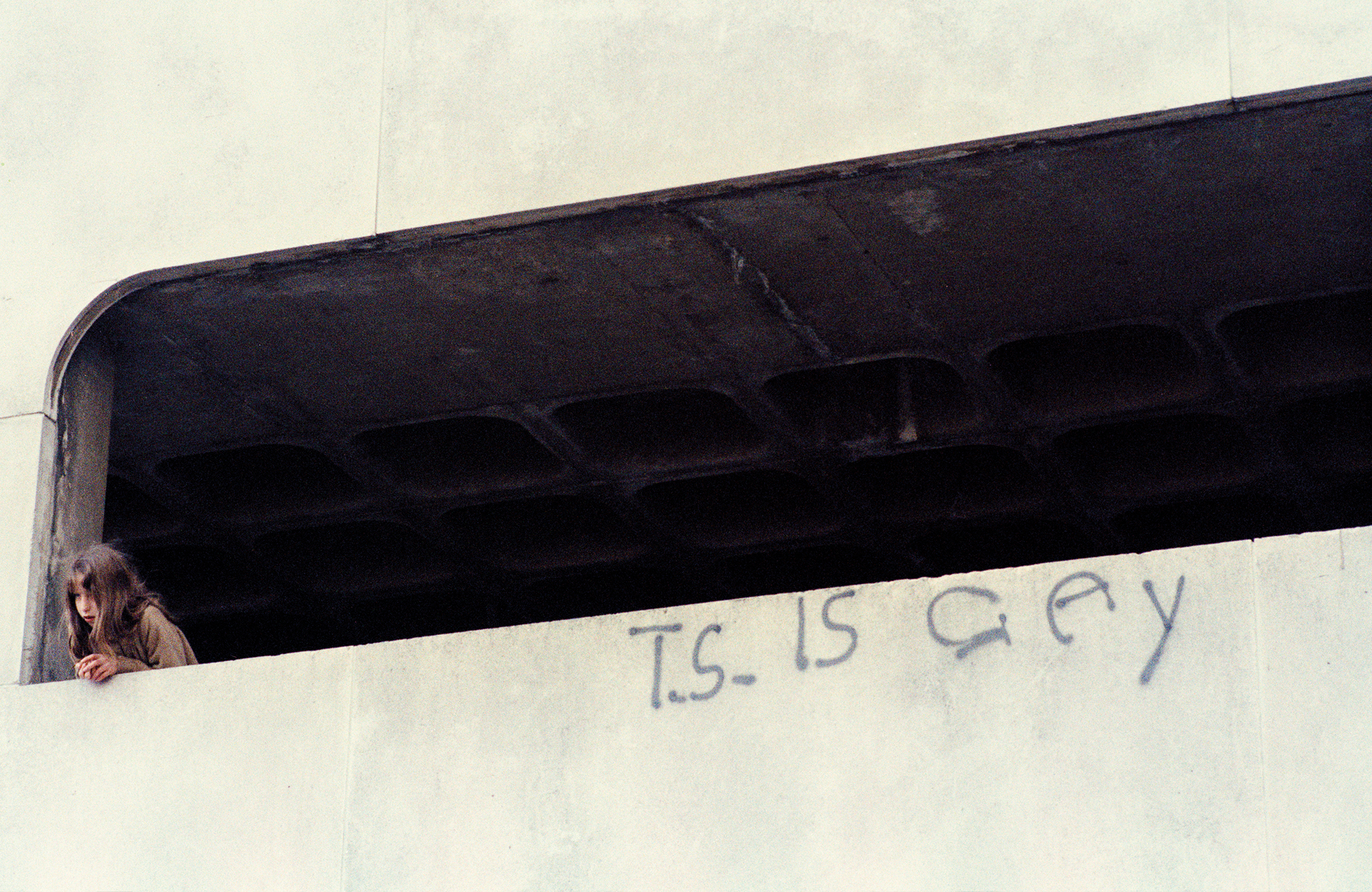 taken by sunil gupta in 80s london, a white girl looks over a balcony with the phrase 't.s. is gay' spraypainted on it