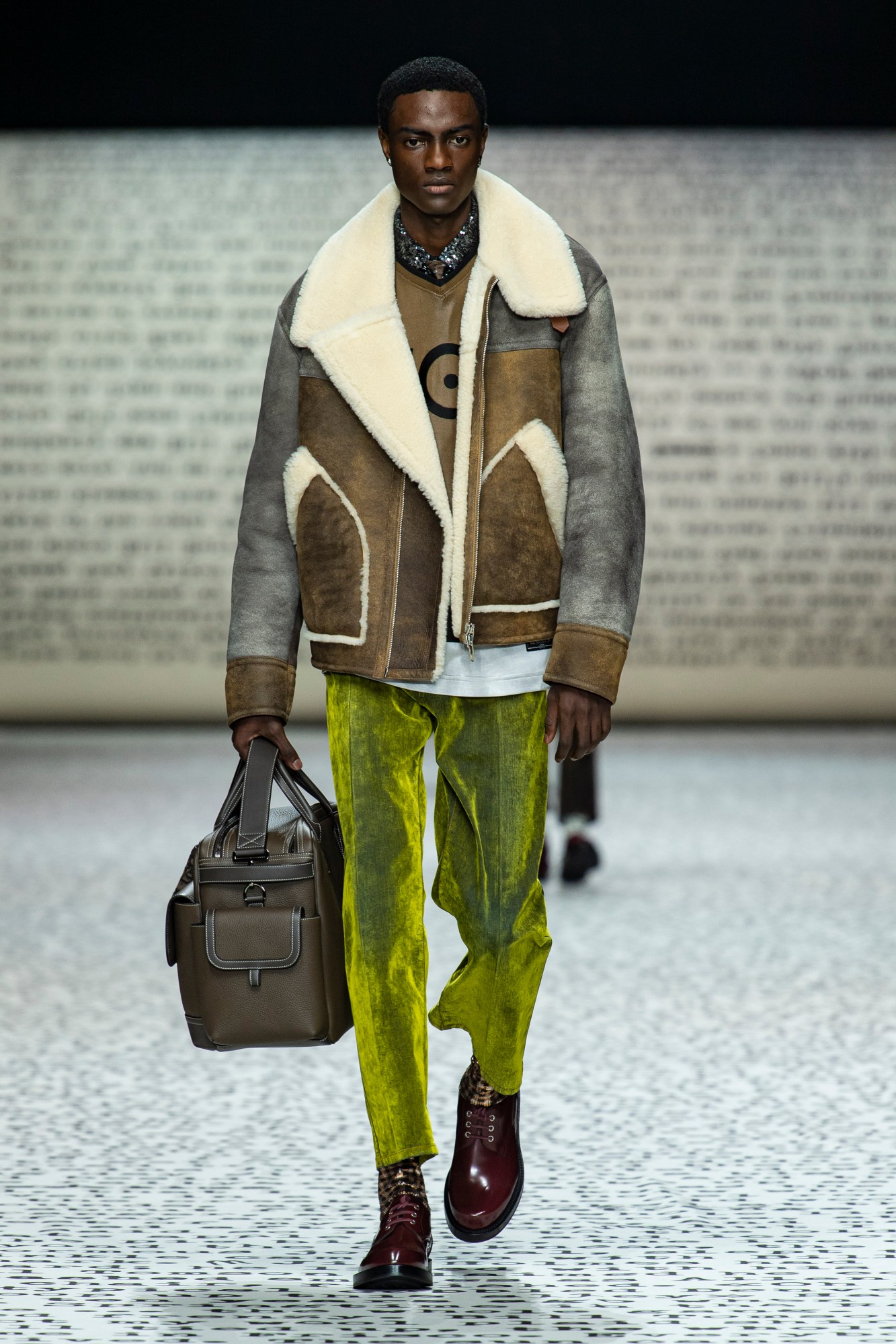 Model wears lime velour trousers with burgundy boots, a blue tee, brown leather sweatshirt and brown and grey shearling lined overcoat with a brown leather bag.