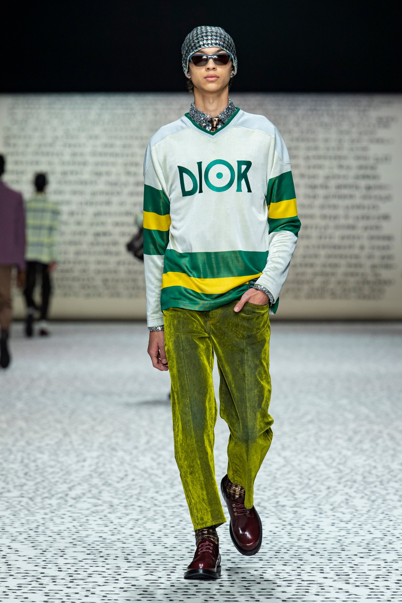 Model wears lime velour trousers with burgundy boots, a floral shirt, gold tie and a Dior sports top over it with a beanie and sunglasses