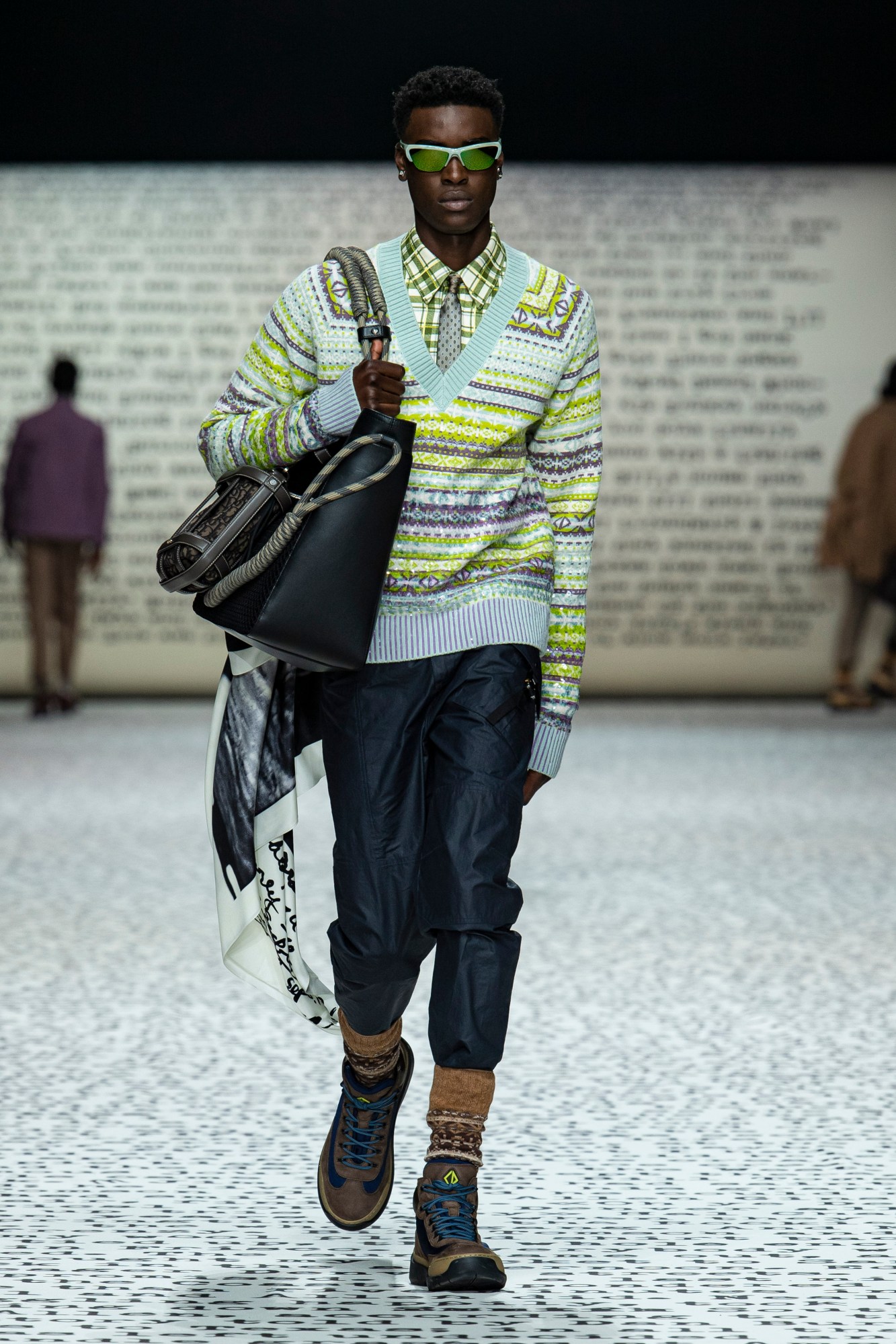 Model wears a green check shirt with a silver tie, green, white and blue fair isle jumper, blue baggy trousers, walking boots and brown socks with a black leather bag.