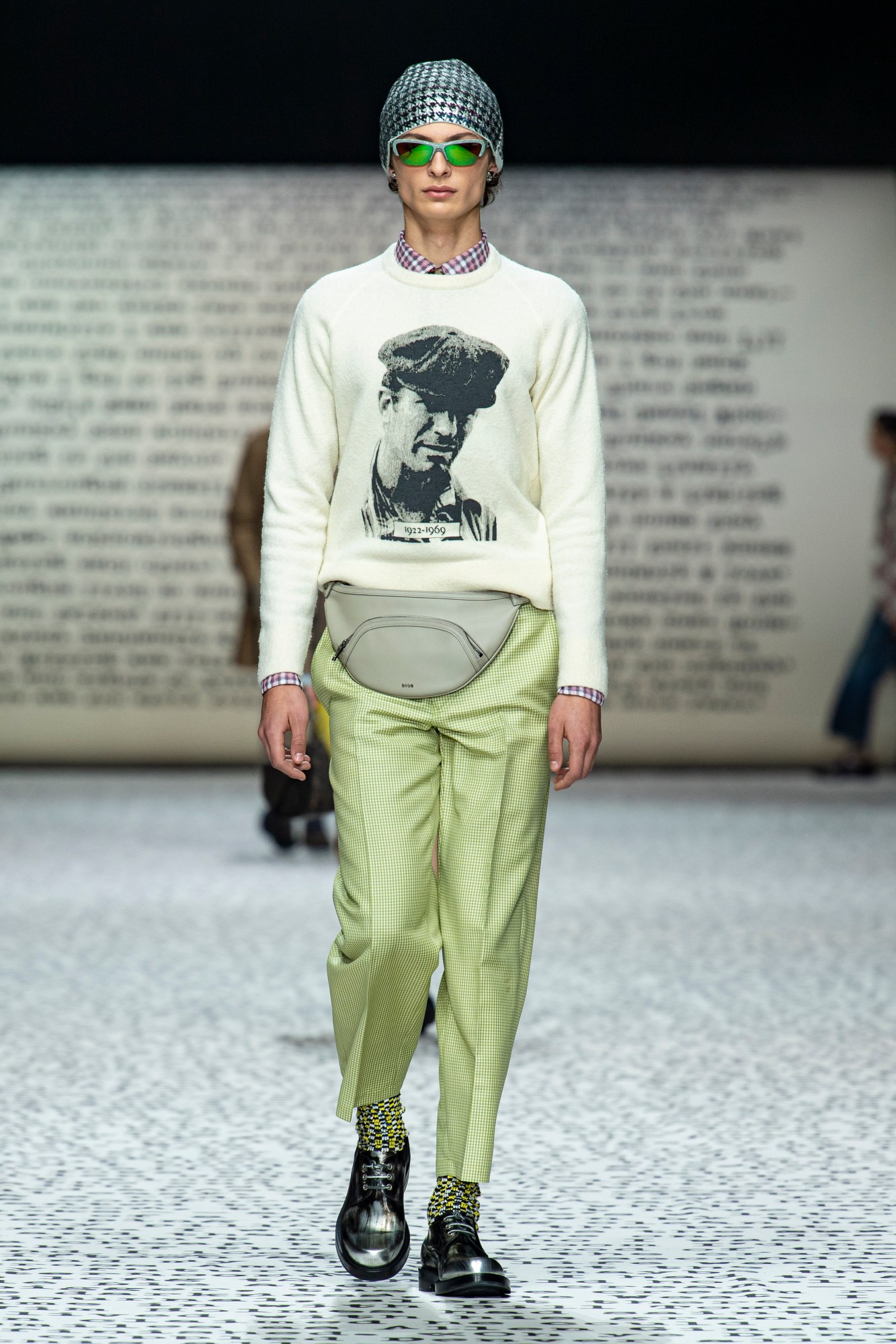 Model wears green trousers with black marble shoes and a printed sweatshirt with a grey bum bag.