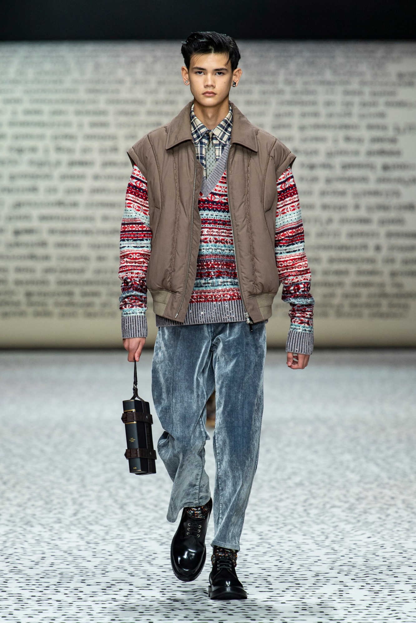 Model wears grey velour trousers with black shoes, a fair isle jumper and a brown gillet.