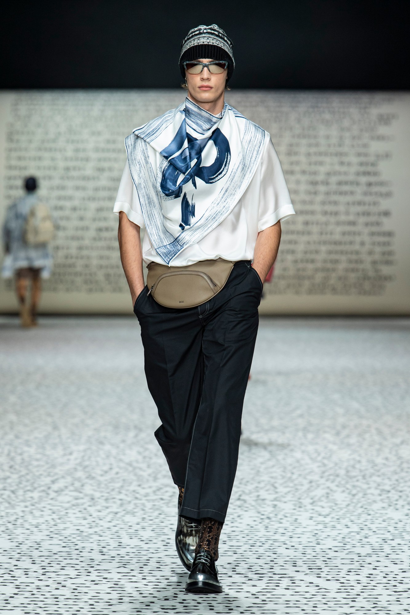 Model wears a printed blue paint white top with black trousers and boots and a bumbag.