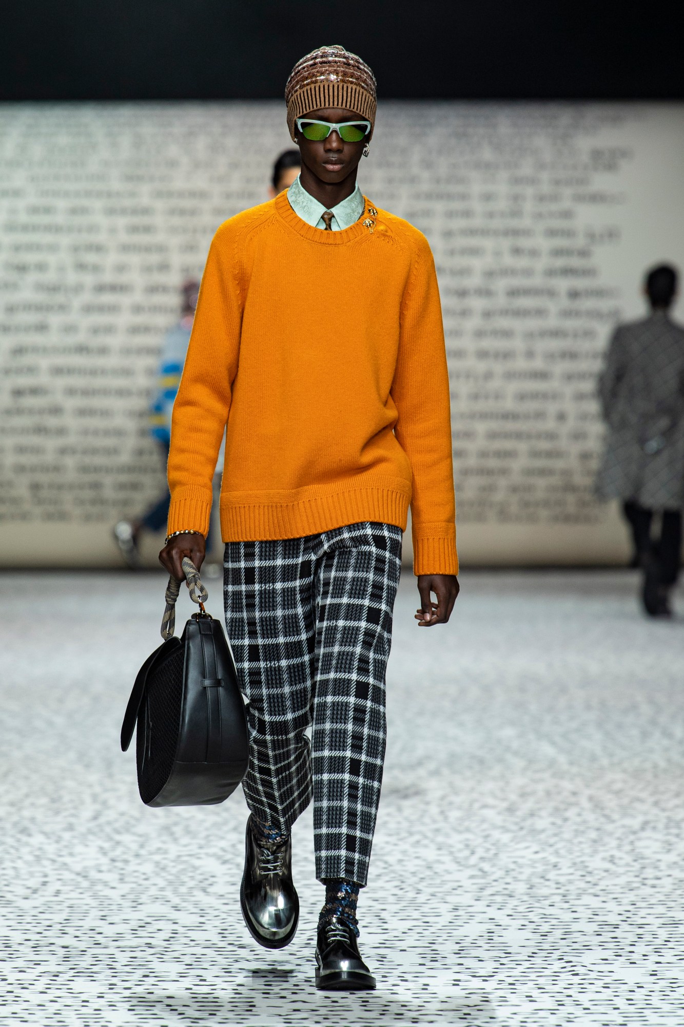 Model wears check trousers with an orange jumper and leather bag.