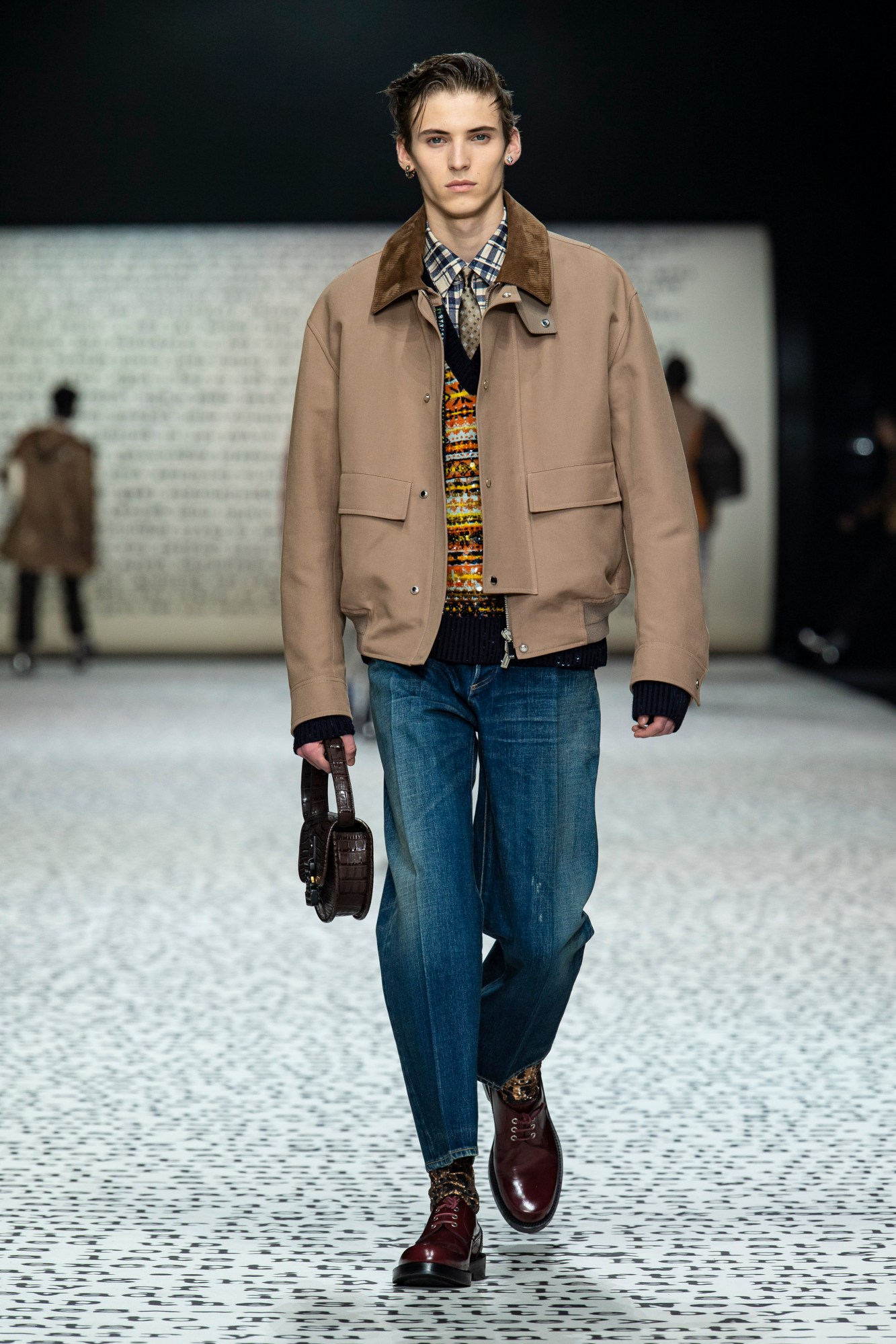 model wears blue jeans with burgundy shoes, a check shirt, fair isle jumper and brown coat.