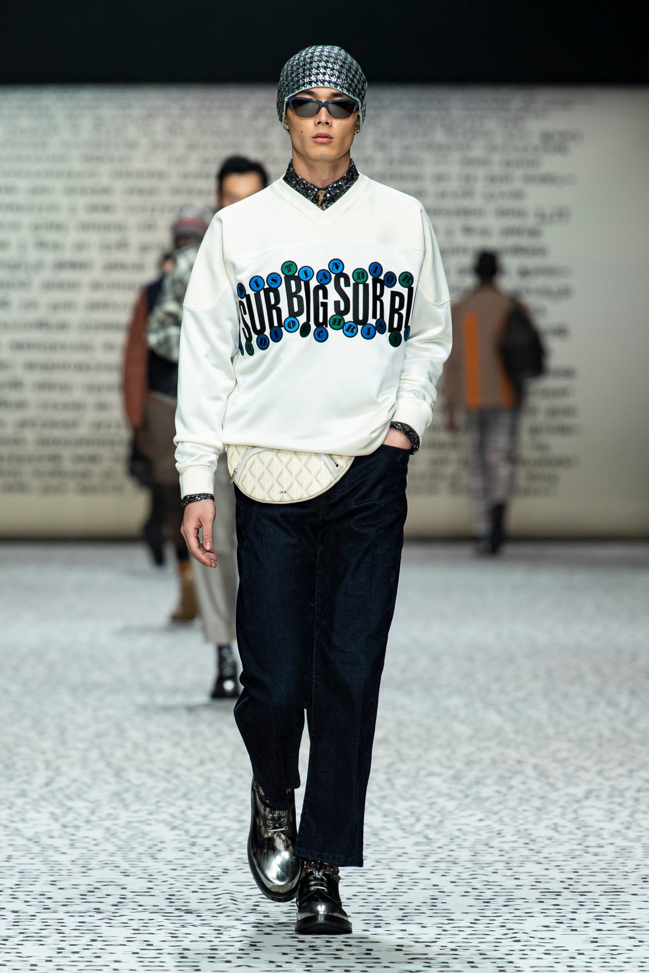 Model wears black trousers with marble shoes, a bum bag and white sweatshirt.