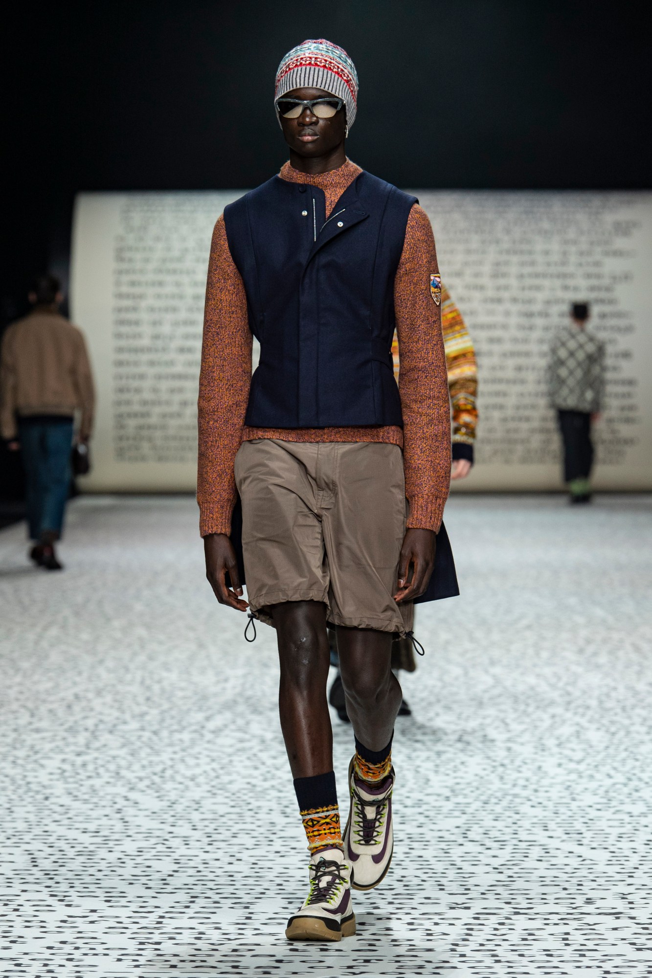 model wears brown cargo pants with white walking boots, an orange turtle neck and blue Gillet.