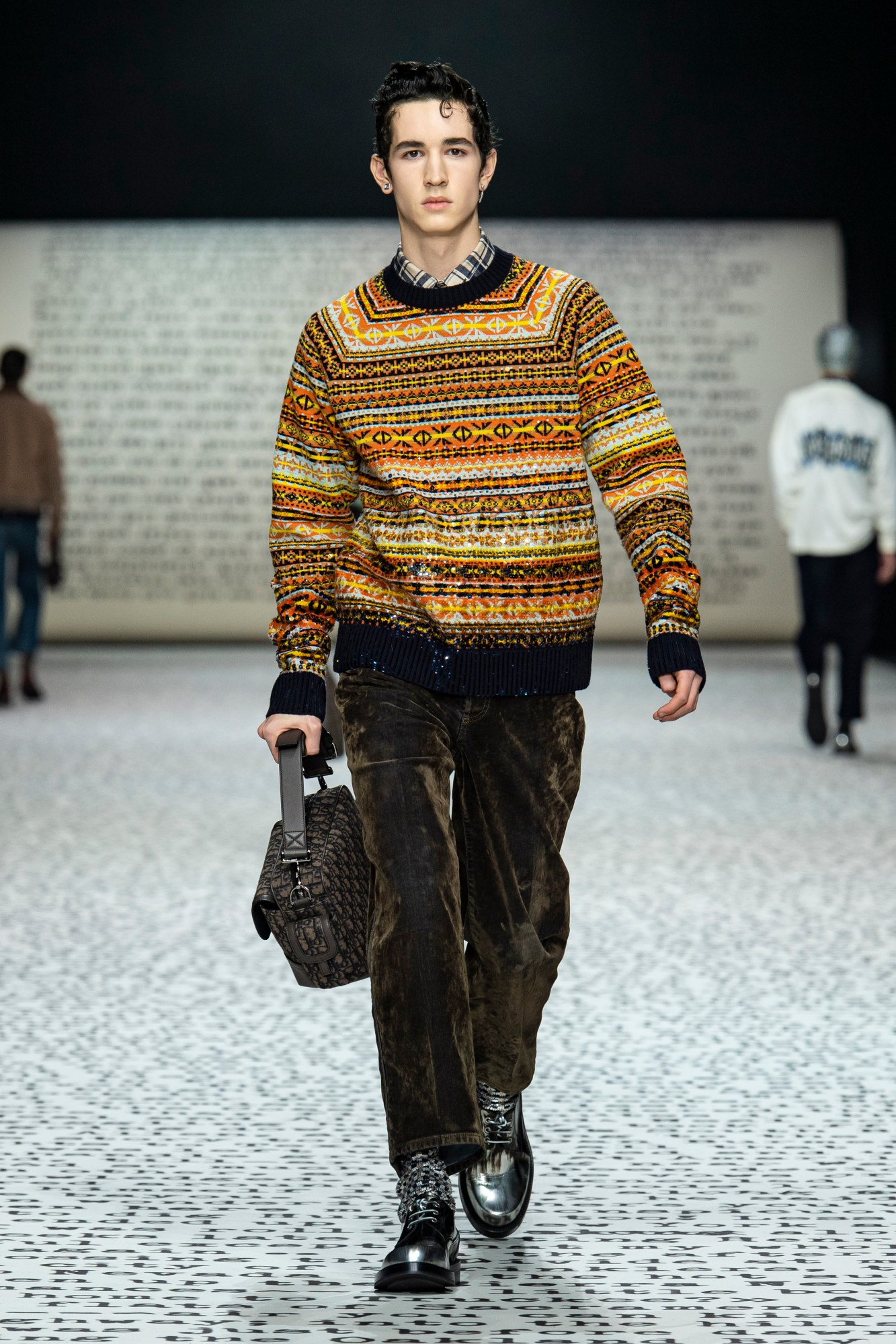 model wears brown velour trousers with a fair isle jumper and a check shirt with a brown bag.