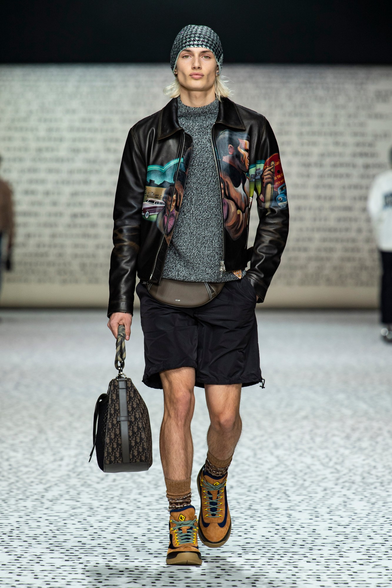 Model wears black cargo shorts with a grey jumper and black leather jacket.