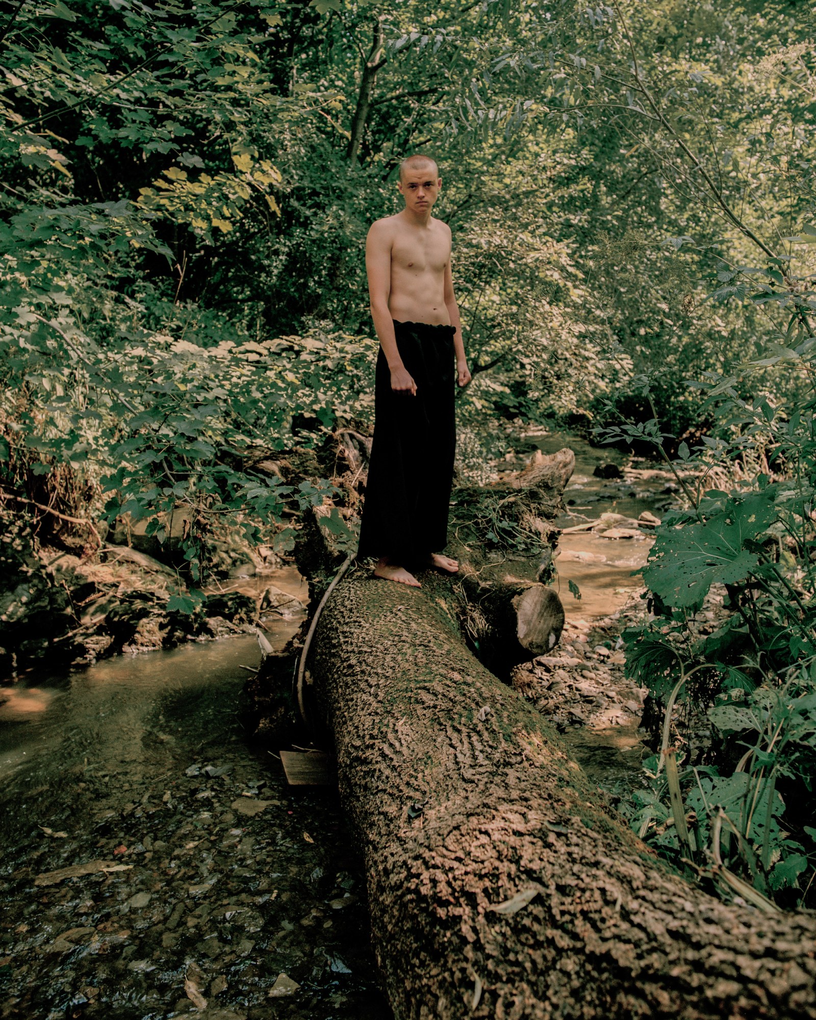 a topless man stood on a tree trunk in the woods