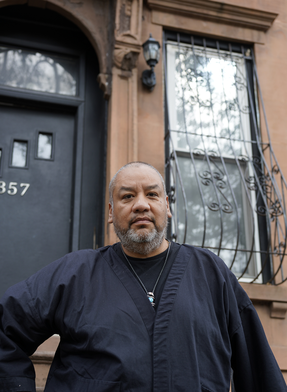 Jeffrey Gibson, wearing black, stands in front of a brown apartment building and its black door.