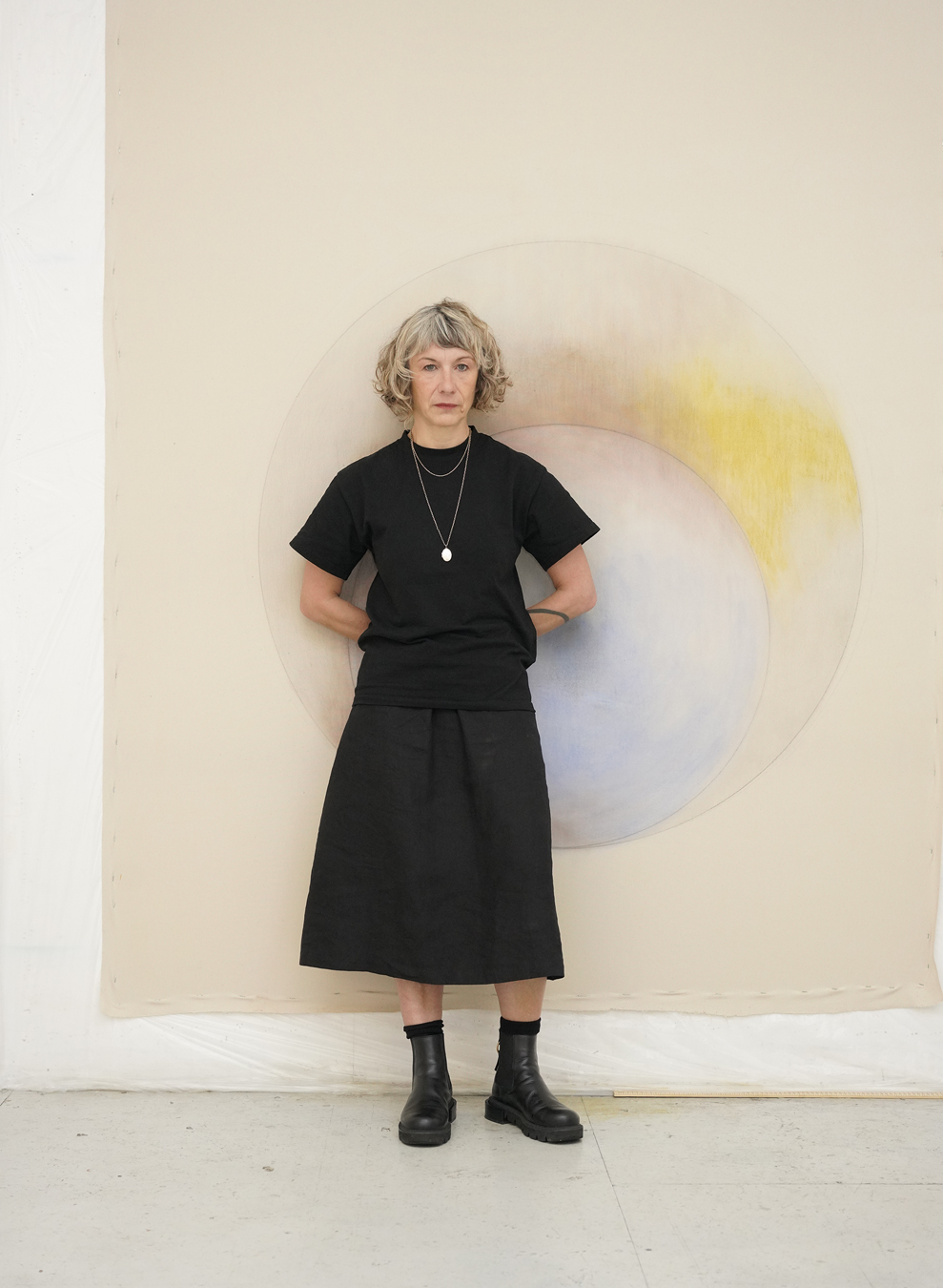 Margarita Gluzberg wearing a black top, skirt and boots stands in front of a spherical painting.