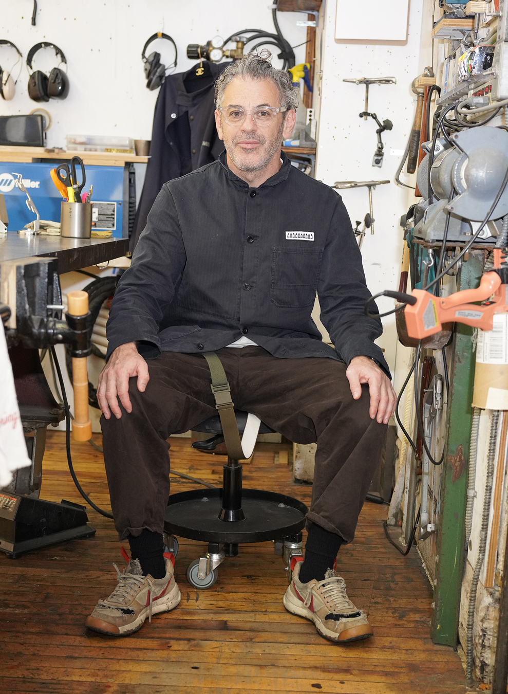 Tom Sachs in a blue shirt, brown trousers and trainers sits in his studio surrounded by tools and art.