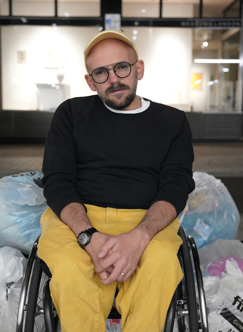 Ryan Gander in a black top and yellow trousers sits in front of trash bags.