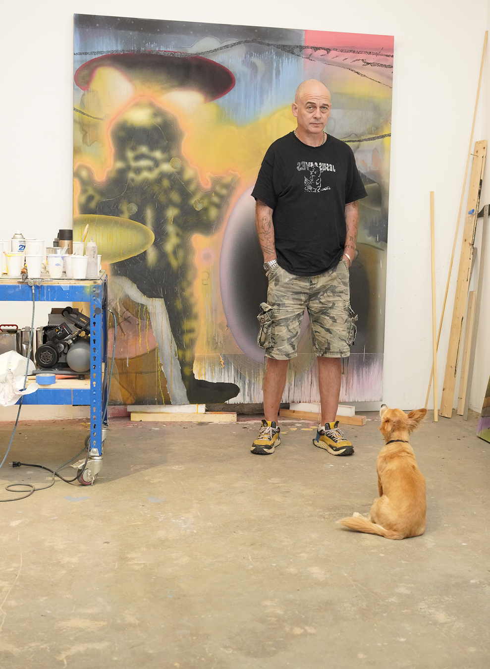 Dinos Chapman in cargo shorts and a black tee and his dog in front of an abstract alien artwork and Dinos tools.