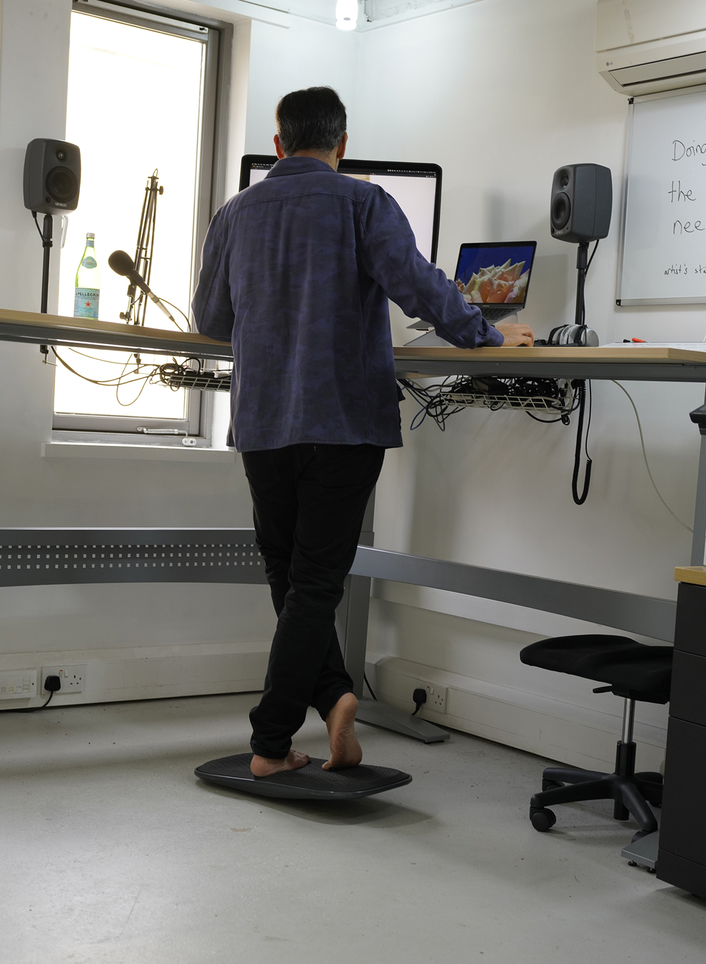 Abbas Zahedi in a blue shirt and black trousers stands barefoot on a balancing mat in front of two computer screens.