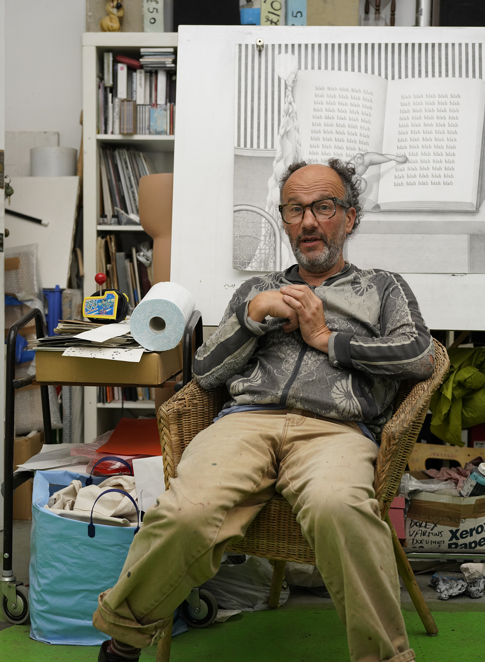 Paul Noble wearing tan chinos and a grey jacket sits in front of his drawing of a book that has the word blah all over it and around the art is books, bags and boxes