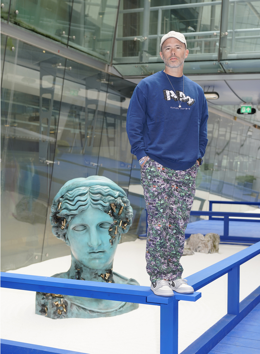 Daniel Arsham in a blue sweatshirt and colourful trousers stand on the frame surrounding his sculpture of the Statue of Liberty destroyed.