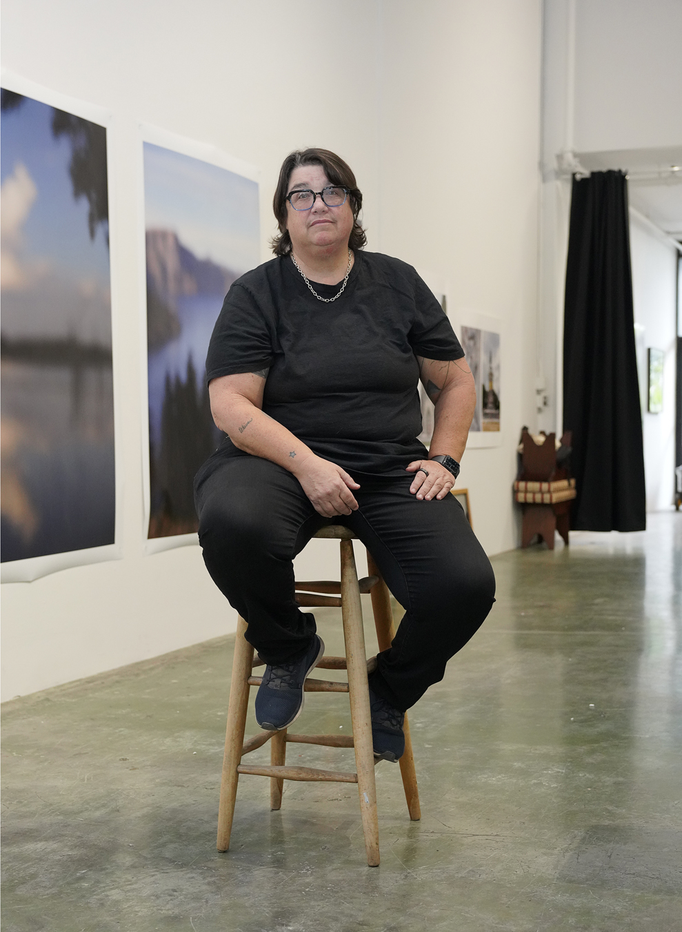 Catherine Opie in all black sits on a stool in front of her art work.