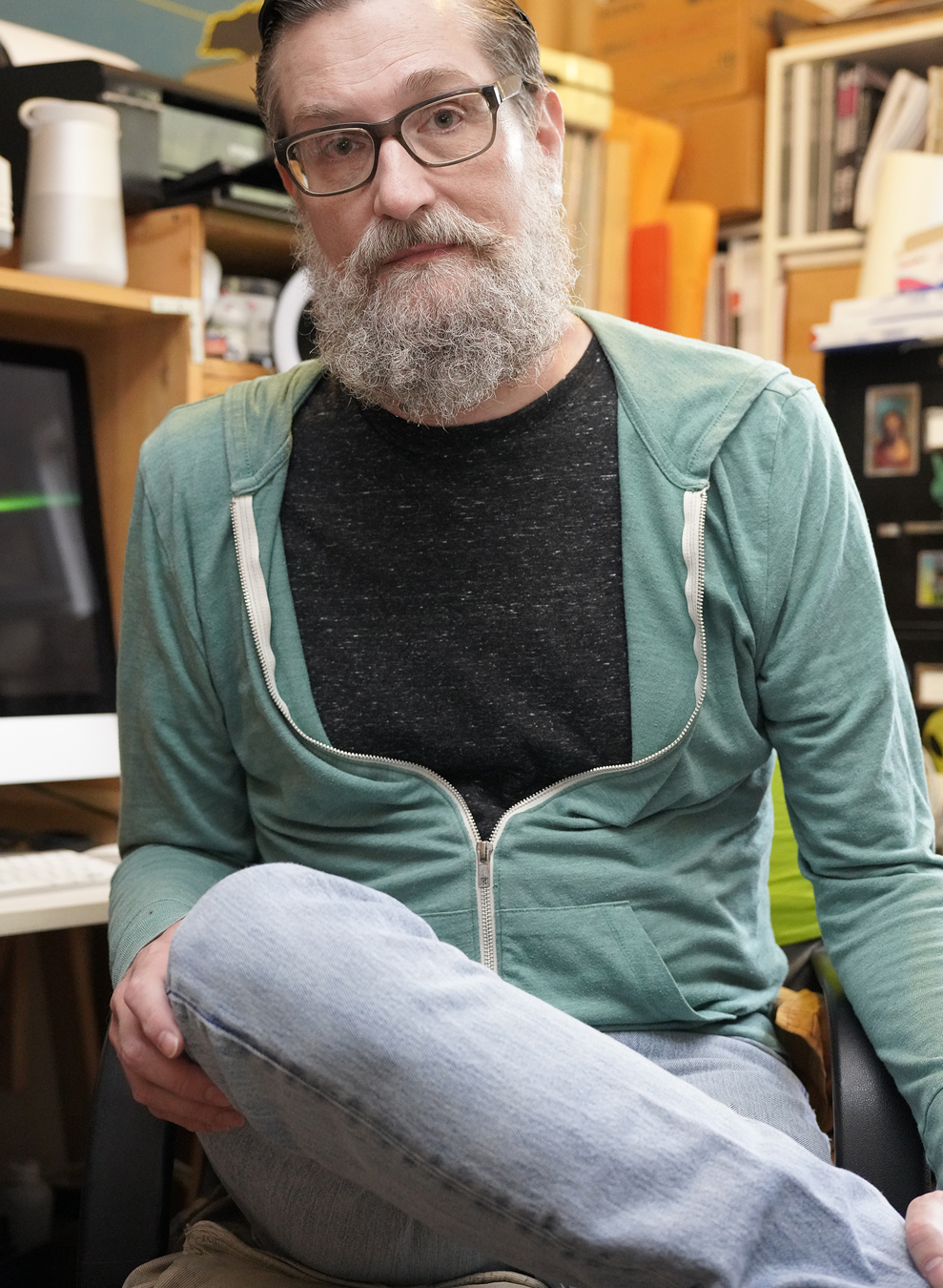 William E. Jones in light jeans and a turquoise hoodie sits in his studio