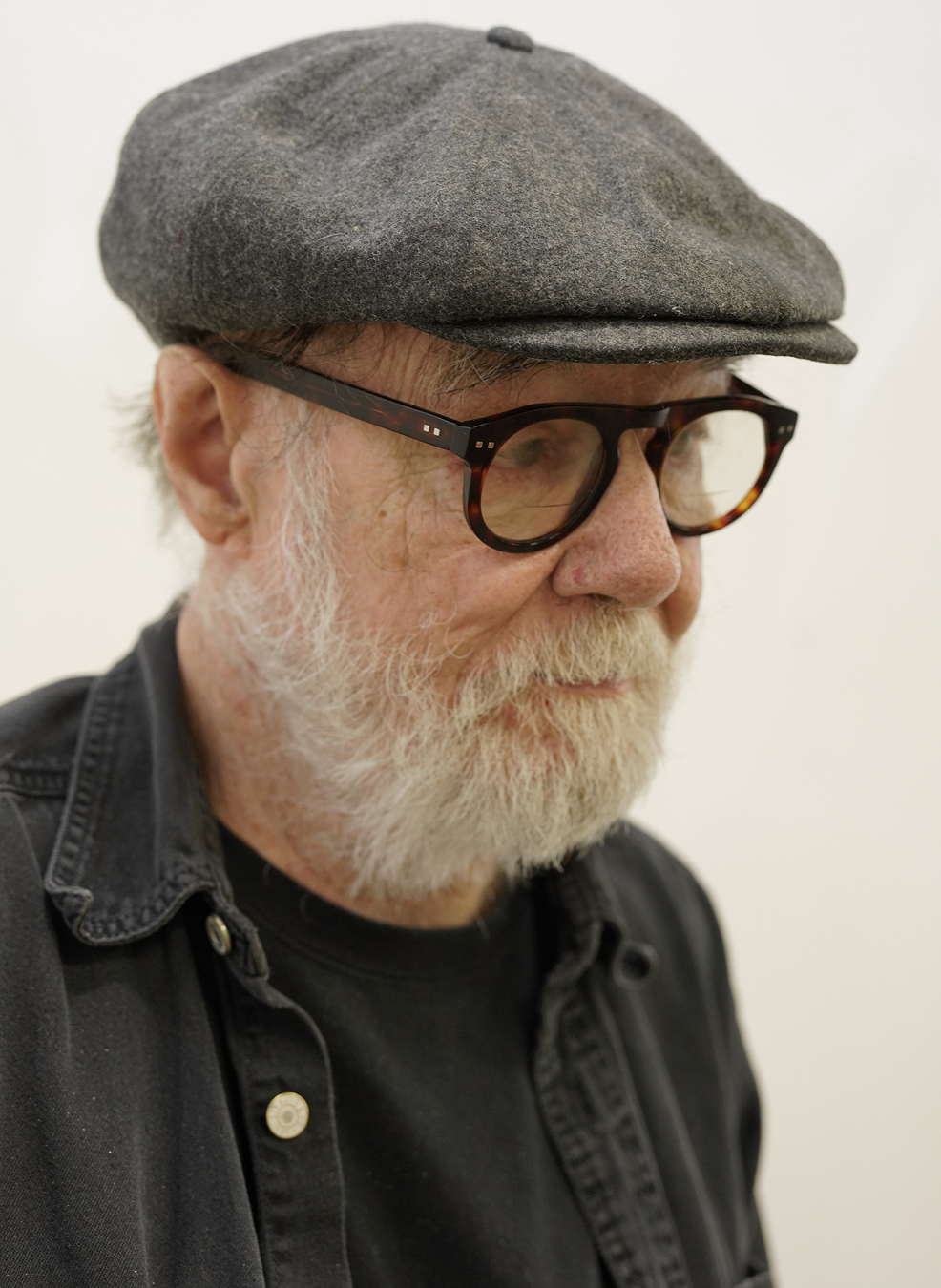 Portrait of Paul McCarthy in a flat cap, a black tee and a black denim shirt.