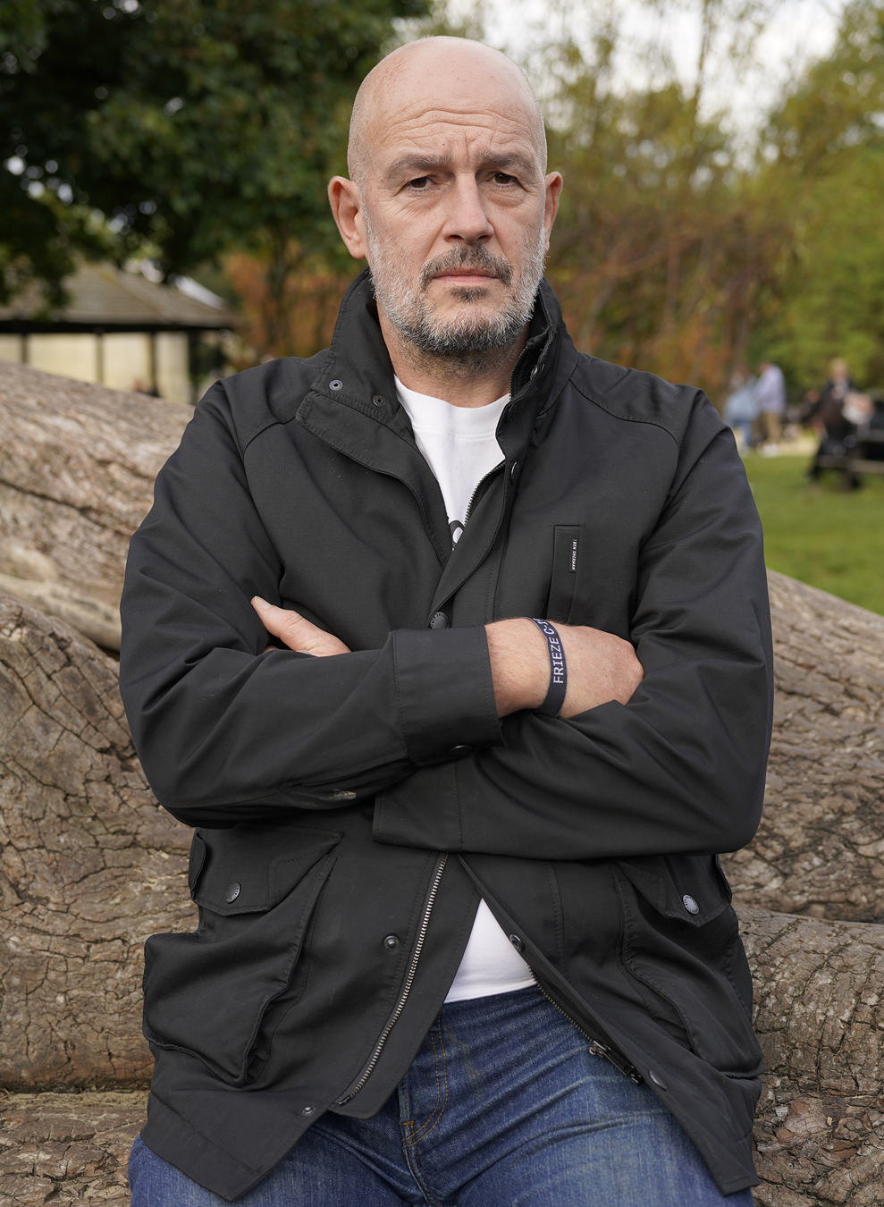 Jake Chapman in jeans, a white tee and black coat sits on a fallen tree.