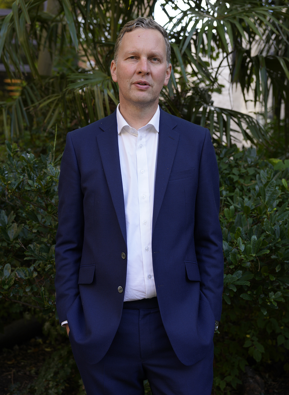 David Shrigley in a royal blue two piece suit standing in front of a bush