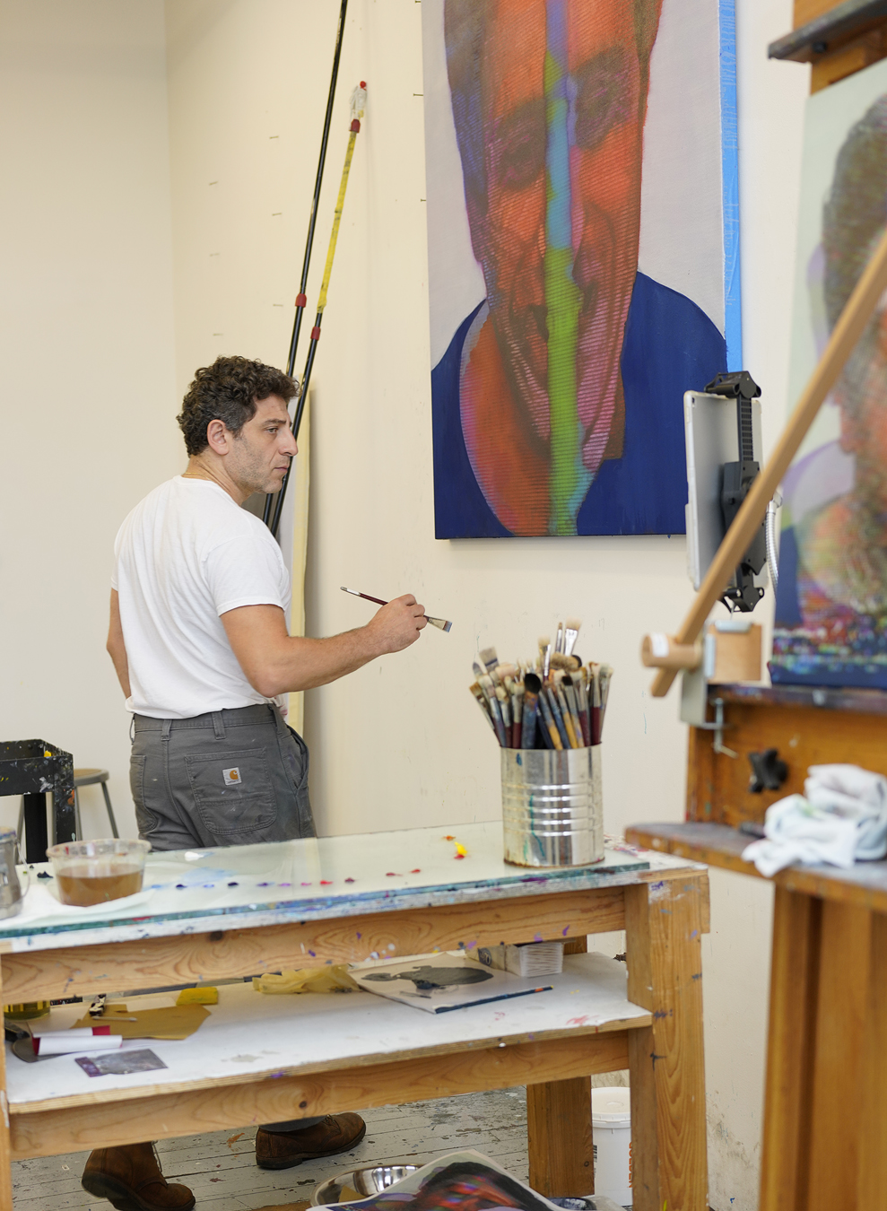 Kon Trubkovich wearing a white top and grey trousers holding a paint brush and standing in his studio in front of a painting and behind an art bench and an easel.