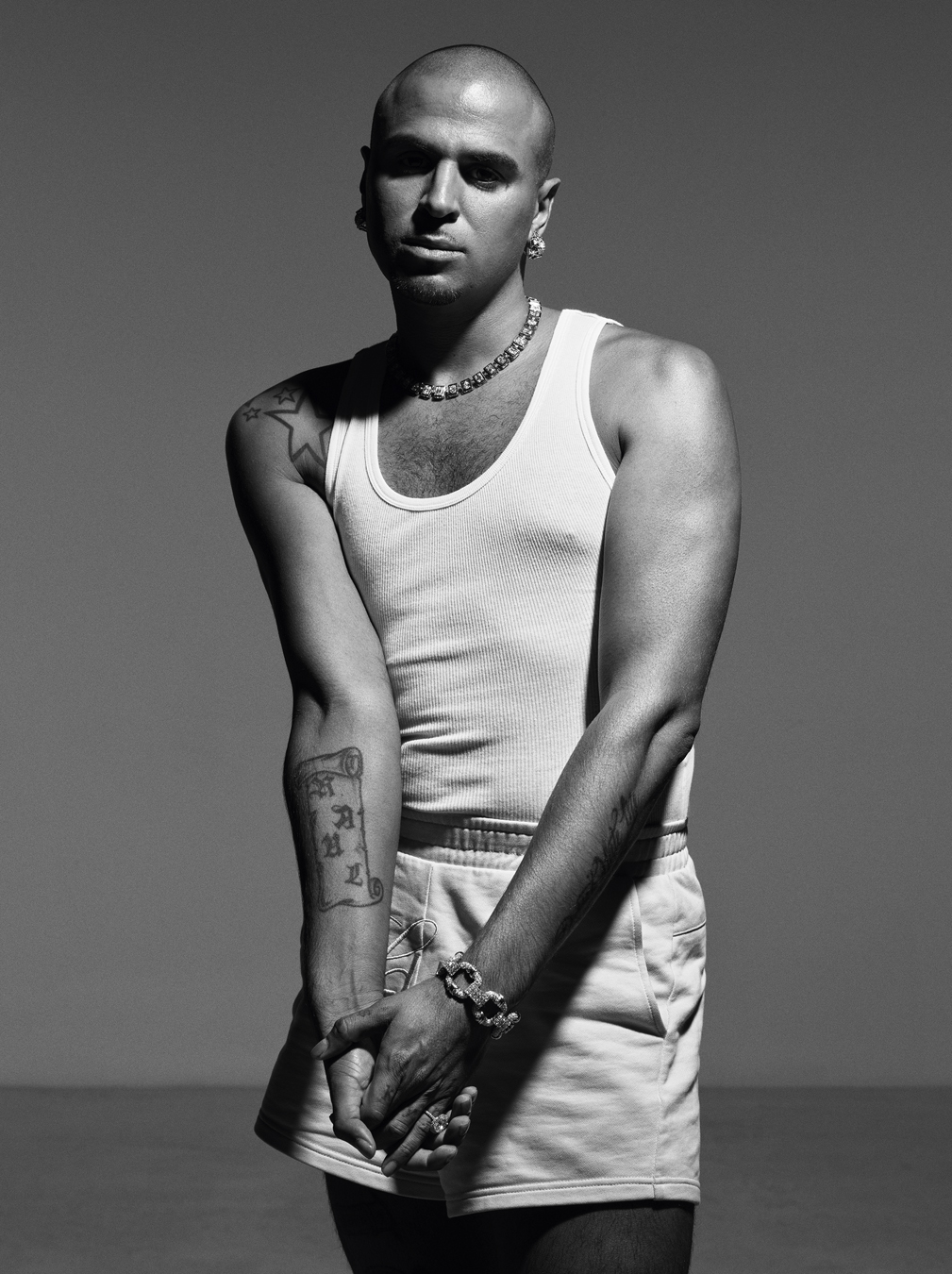 raul lopez stands wearing shorts and a vest top, with a jewelled necklace and bracelet. his hands are clasped in front of him