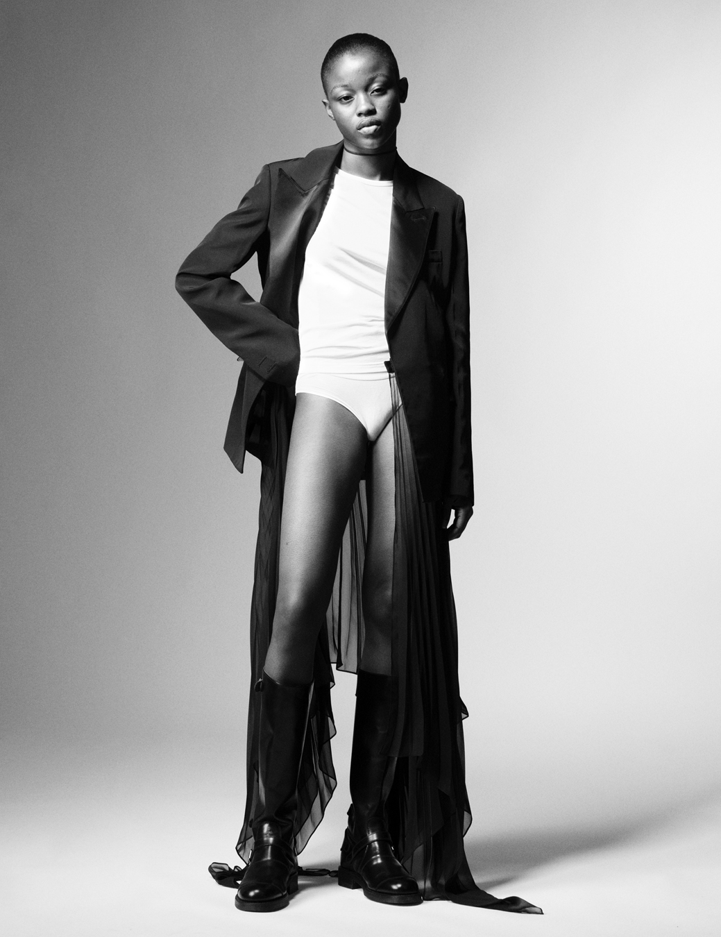 Model wears a white vest and briefs with a black blazer and sheer sarong skirt with boots.