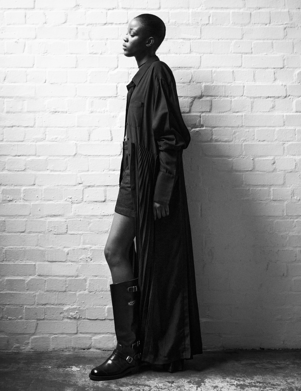 Model stands against a brick wall wearing a black coat and black shorts and black boots.