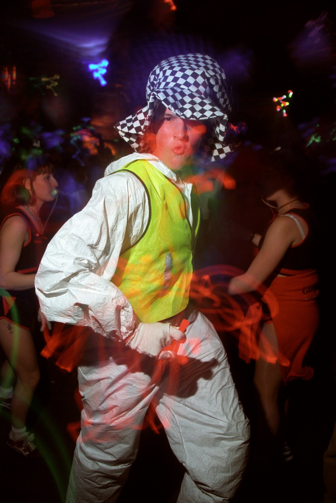 a man in a white boiler suit, high vis vest and checkered hat dances at a rave in the 90s