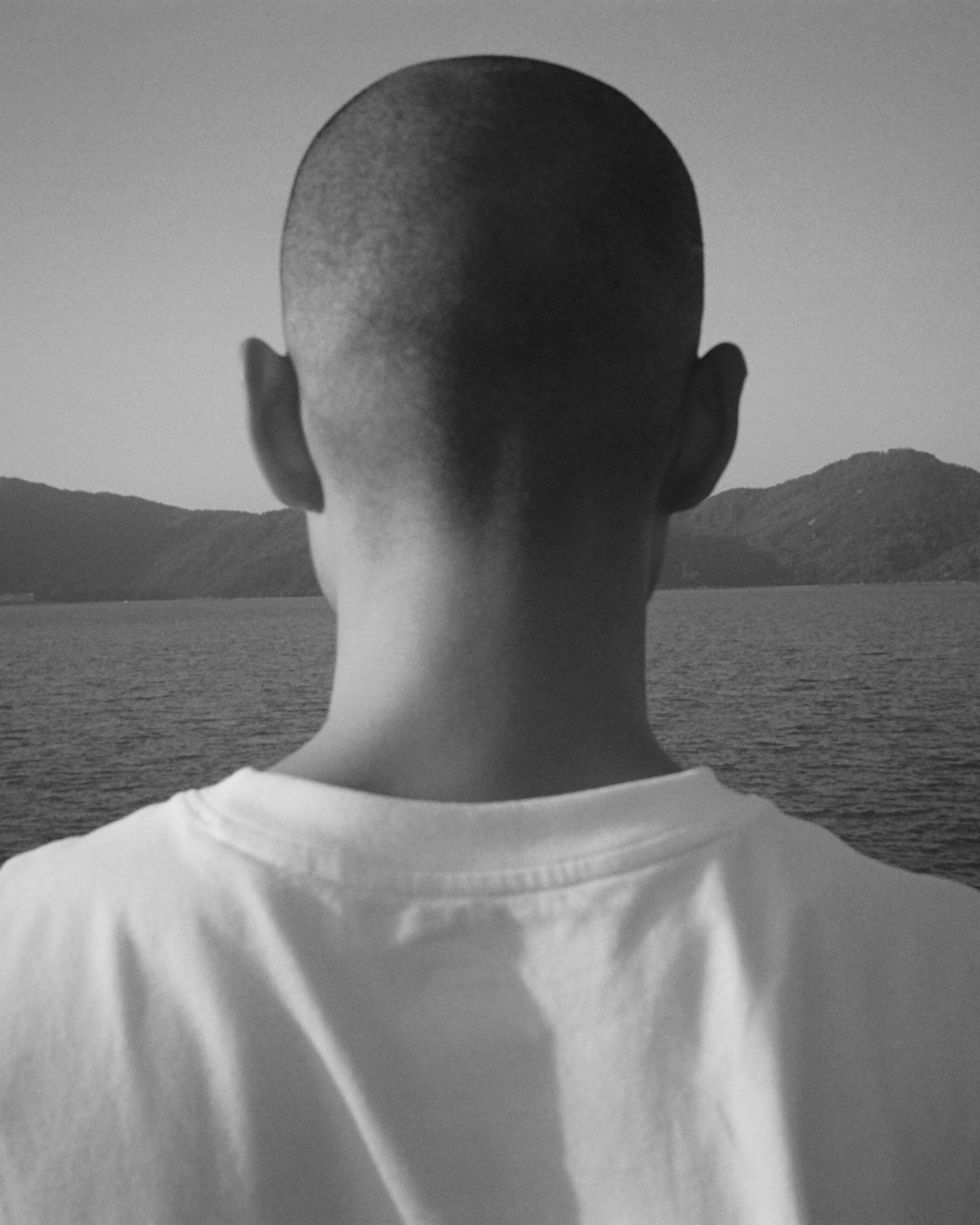 The back of the shaved head of a man in a white t-shirt looking out over a lake and mountains.