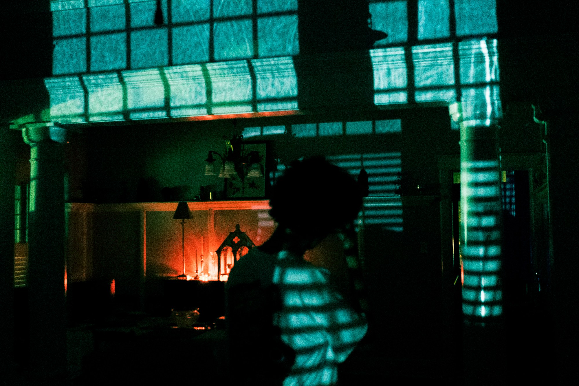 A man in a dark room stands in front of a wall and lit up font. The light and rain from between the window blinds reflects onto him.