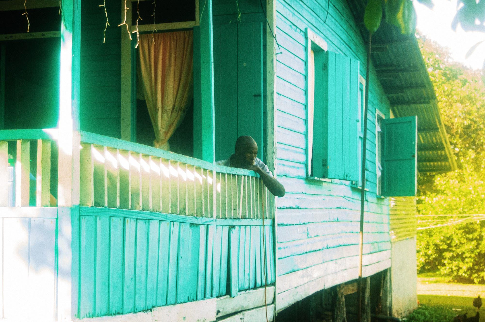 A man leans over his porch decking fence of his wooden blue house, looking into the garden.