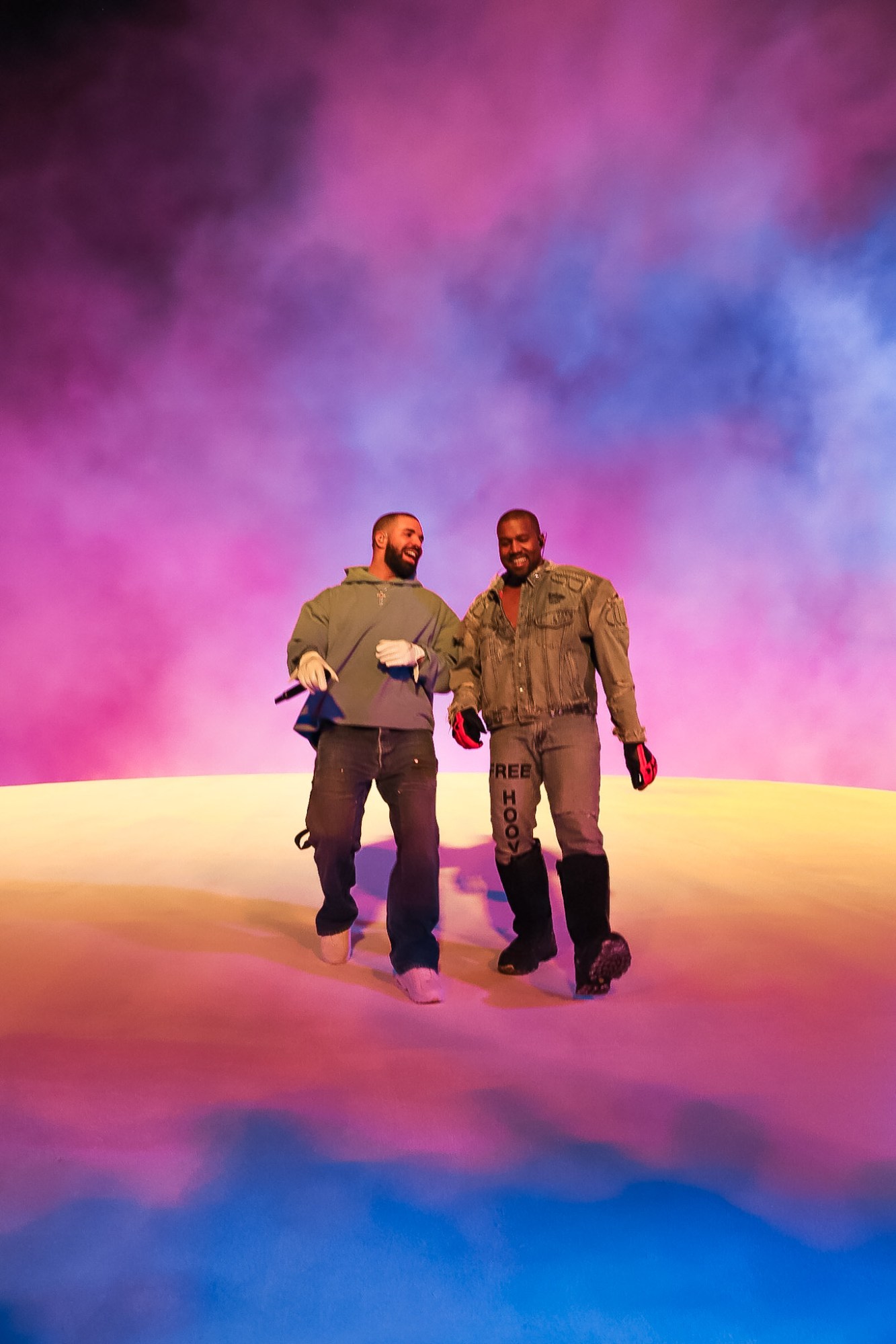 Drake and Kanye West walking along together across a rainbow coloured floor and a pink smoky sky.