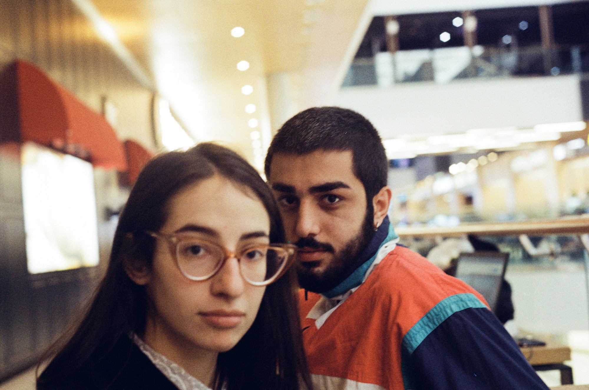 A man with short hair and a sports jacket and a woman with retro 70s glasses and long straight hair look at the camera as they stand in an office building.