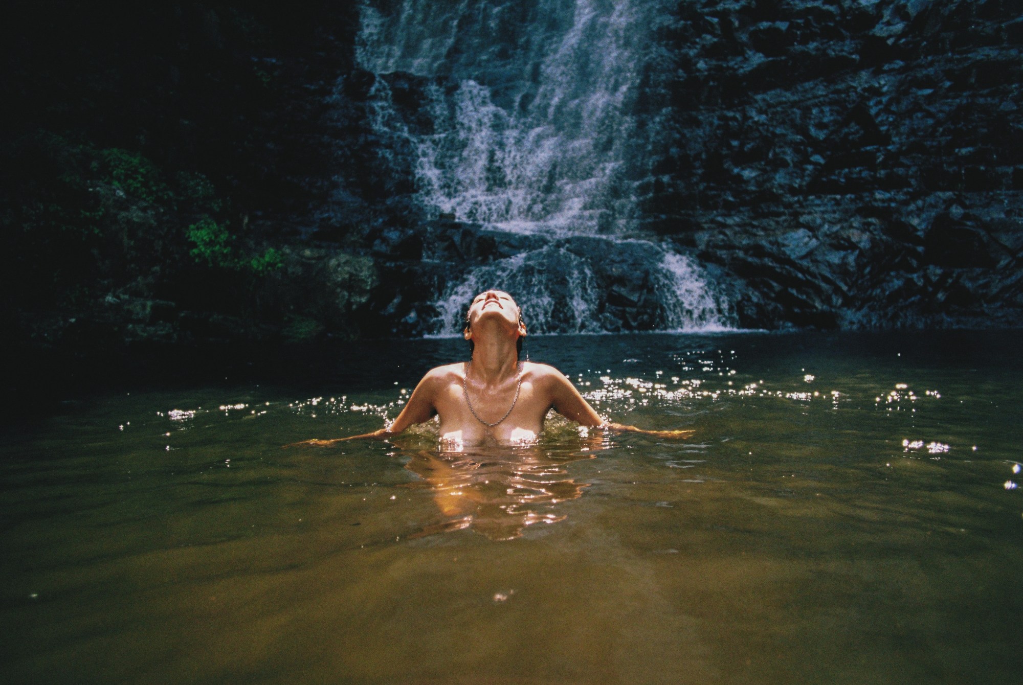 A naked woman looks up at the sun as she swims in a lake with a waterfall in the background.