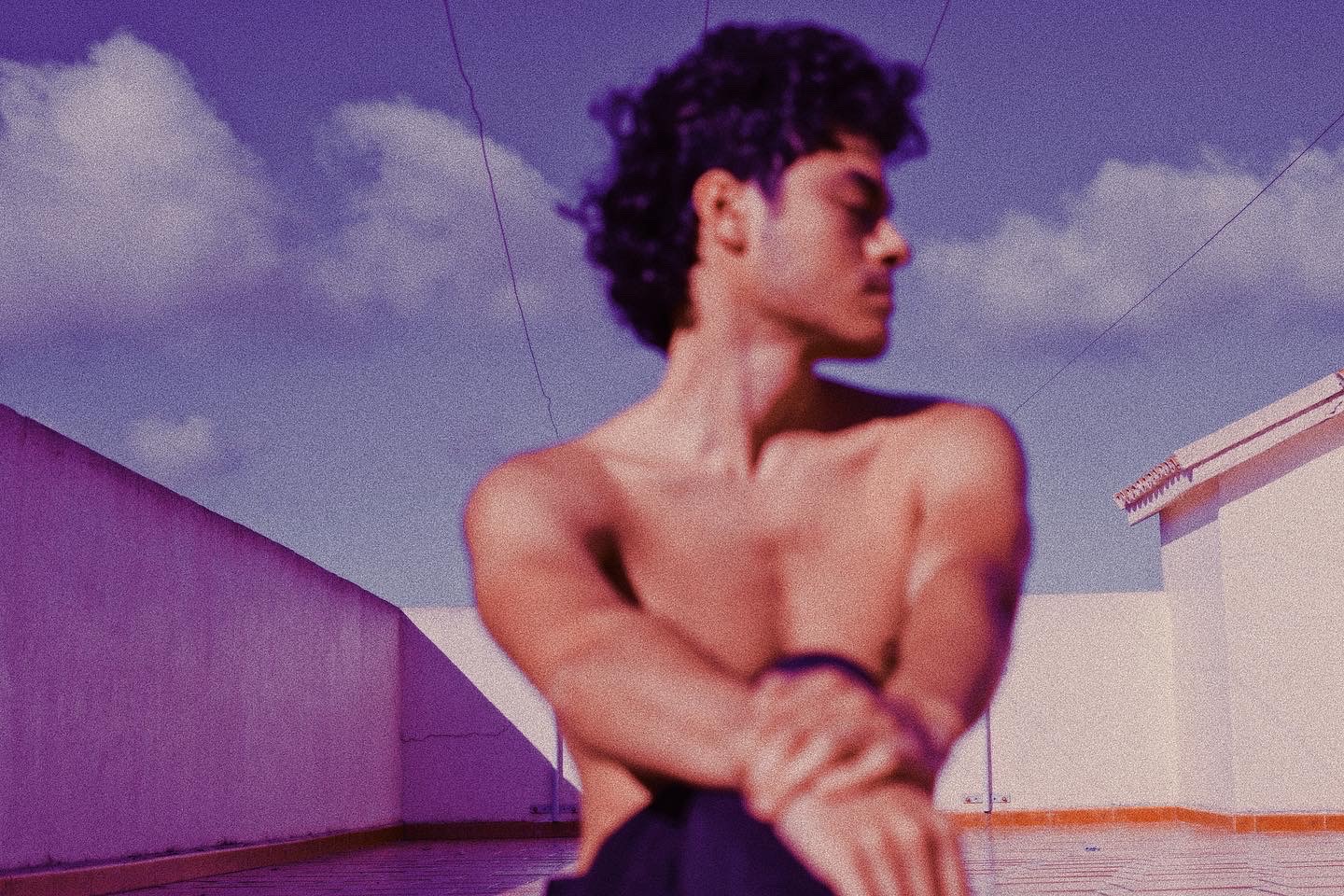 A man sits shirtless on a roof, his hands meeting on his knee as he stares off to the right.