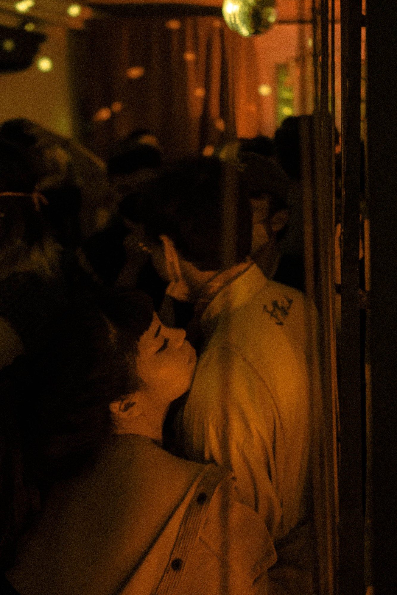 An individual with a corduroy jacket and long tied back black hair leans on the shoulder of a man in a white shirt amongst a crowd of people under strobe lights and a disco ball.