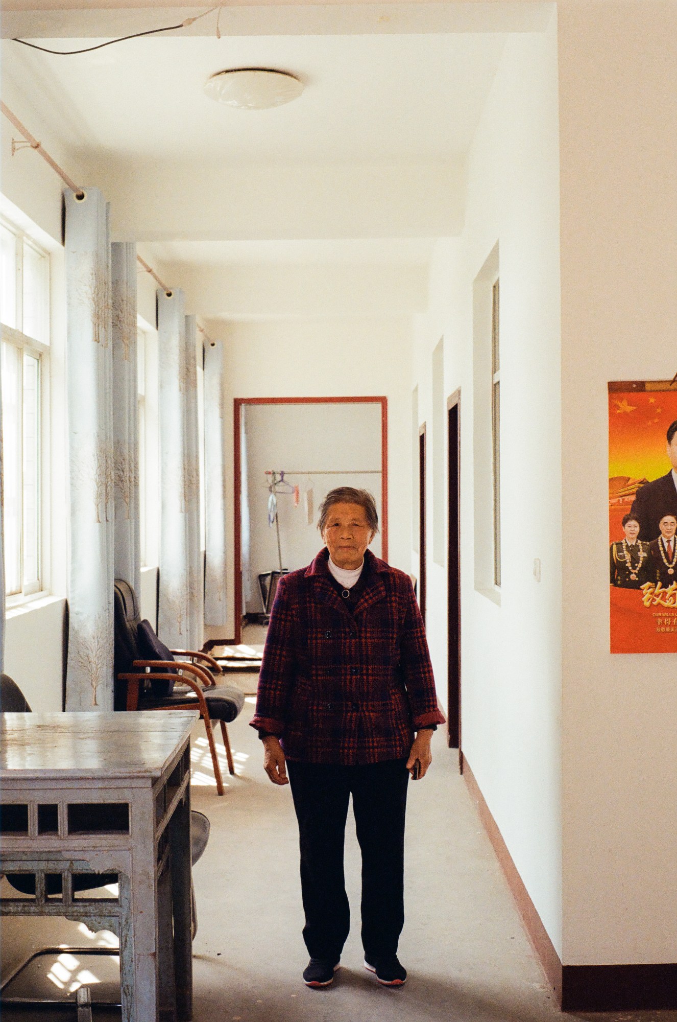 An old lady in a red tartan jacket and black trousers stands in a corridor. Next to her is a table and chairs and beside her is a poster.