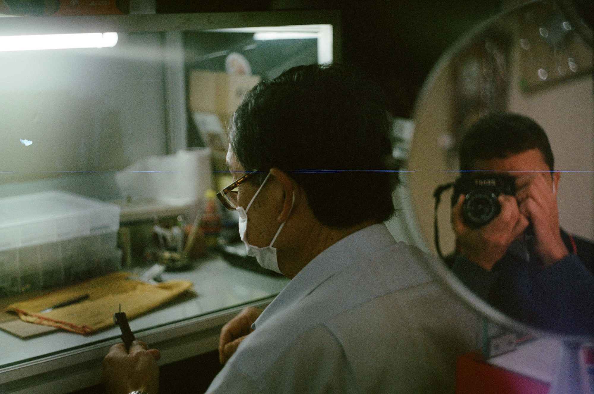 A man wearing a shirt and mask sits at a desk as the photographer shooting him can be seen reflected in a mirror next to him.