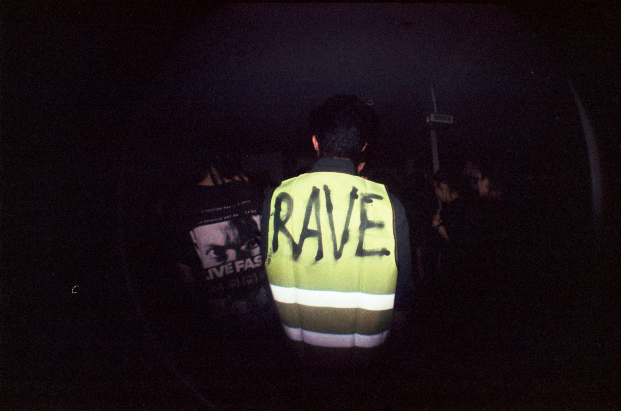 The back of a man outside at night wearing a high visibility jacket that has RAVE spray painted on the back.