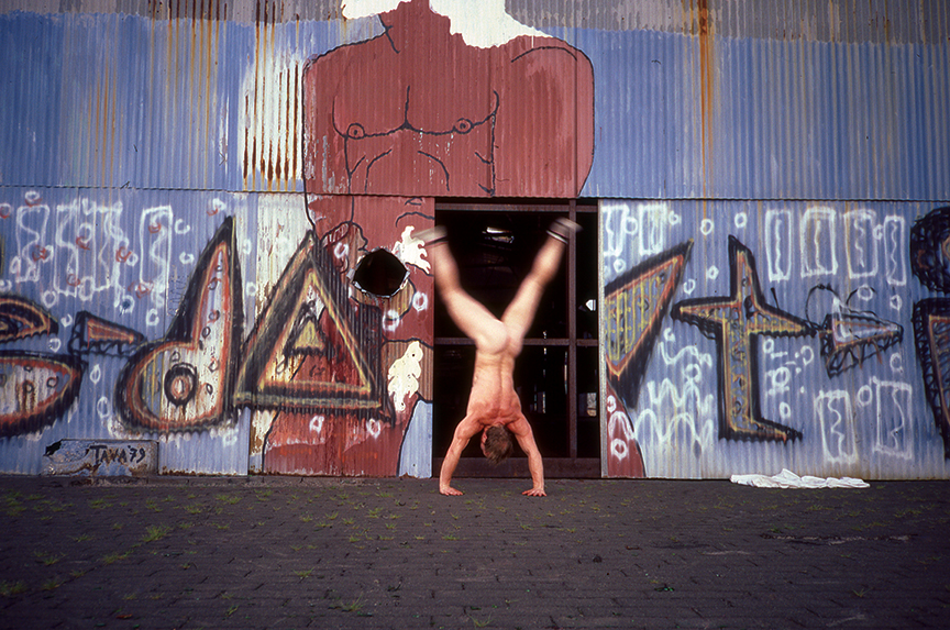 a naked man doing a cartwheel in front of graffiti at christopher street pier