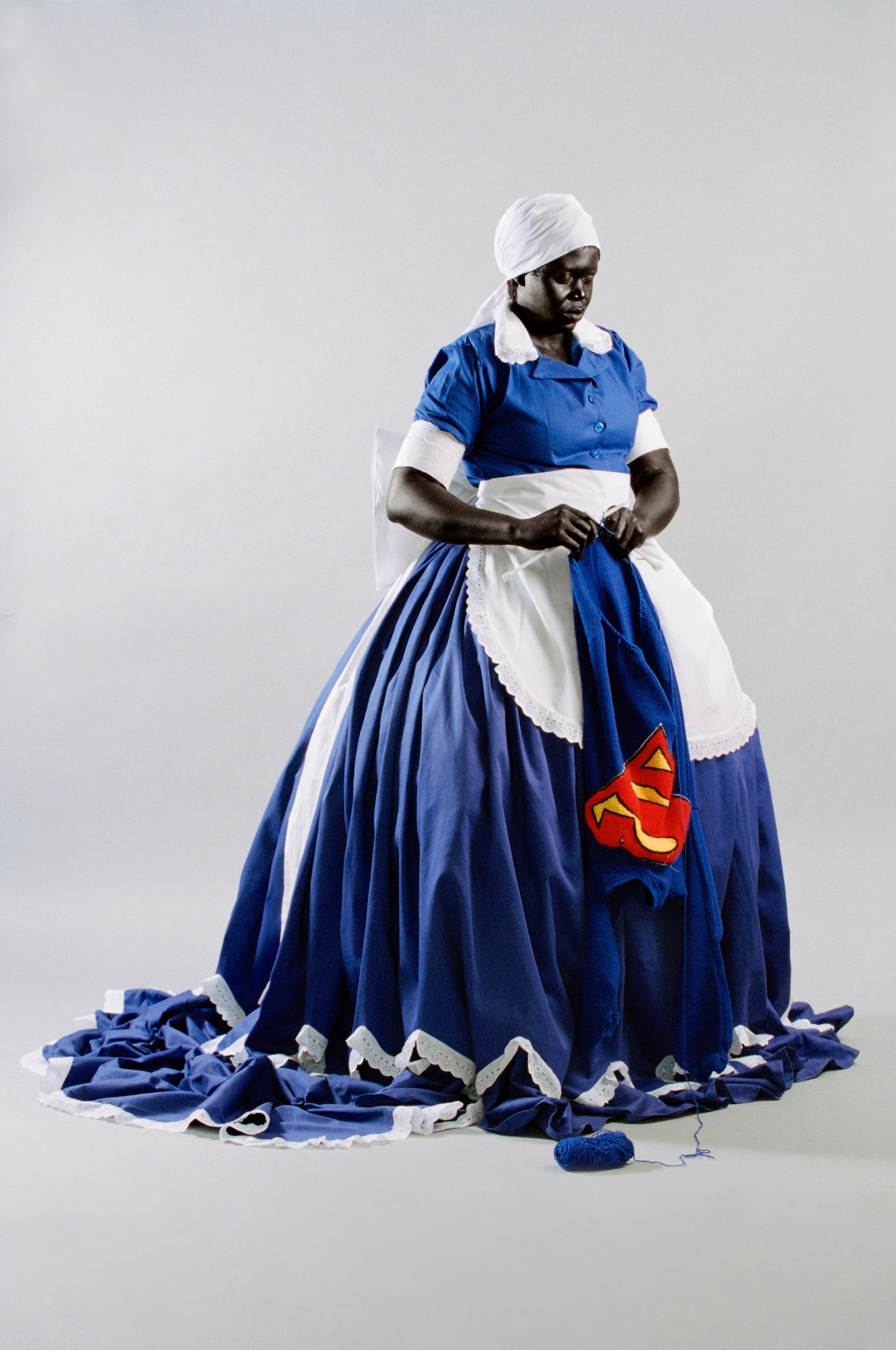 a black woman in a modified maid's dress sews a blue sweatshirt bearing a superman 's'